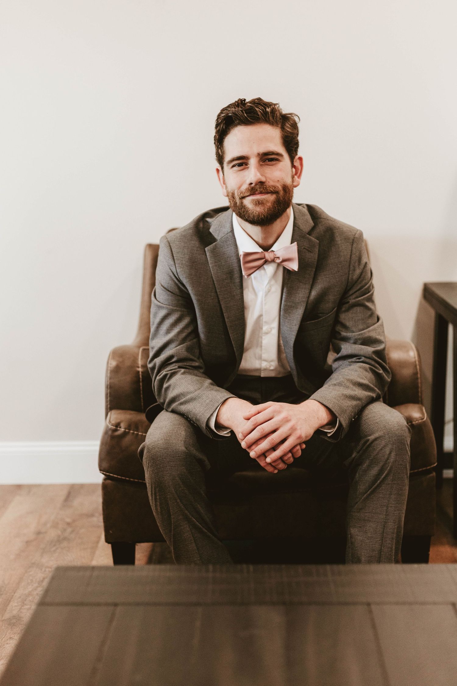 Handsome groom in gray suit and pink bow tie sitting in leather chair smiling.