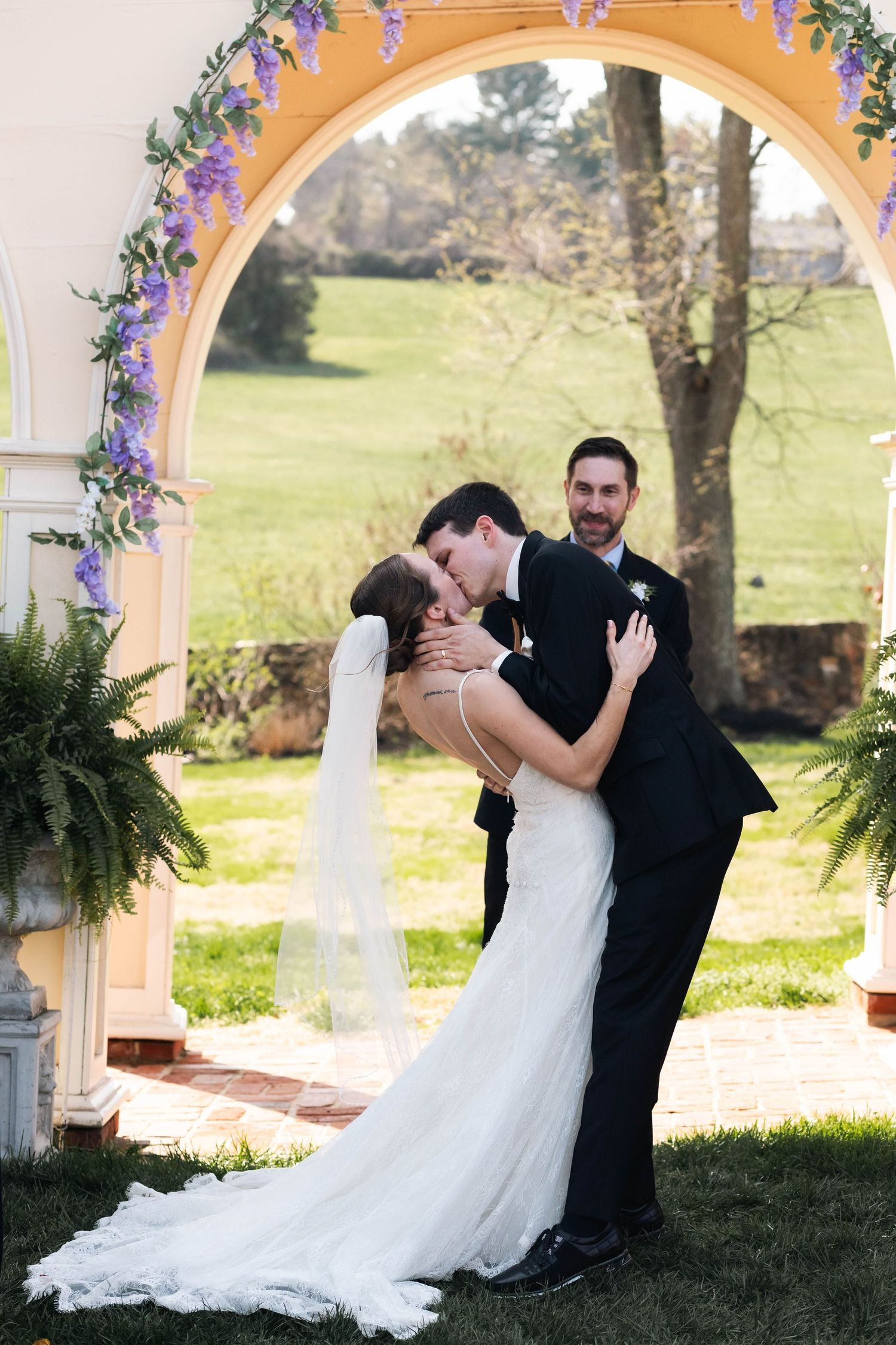 Bride and groom share first kiss under a yellow arch adorned with purple flowers at outdoor wedding ceremony.