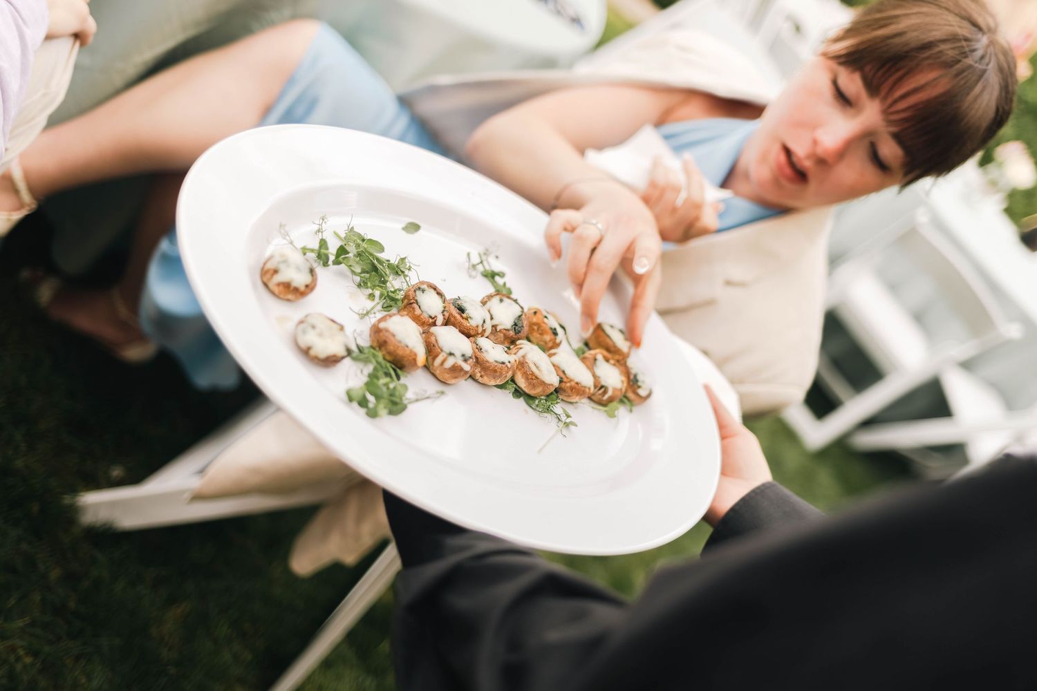 Server presenting elegant platter of gourmet appetizers with microgreens to seated wedding guests.