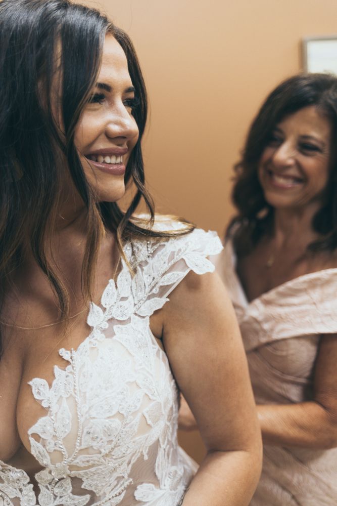Radiant bride in lace wedding gown smiles joyfully while getting ready with loved ones.