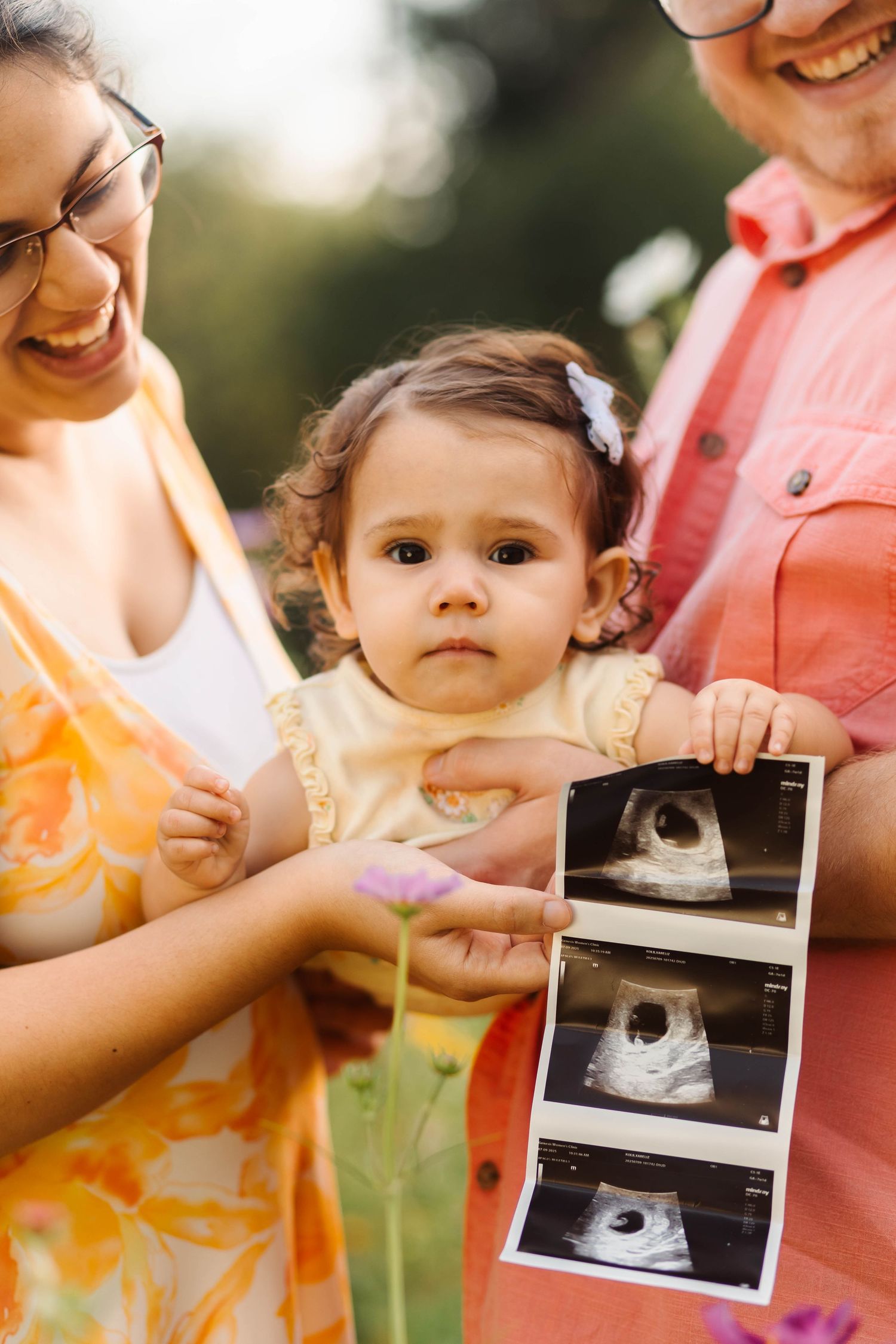 Smiling parents with toddler girl holding ultrasound photos in sunny garden in Perkiomenville, PA.