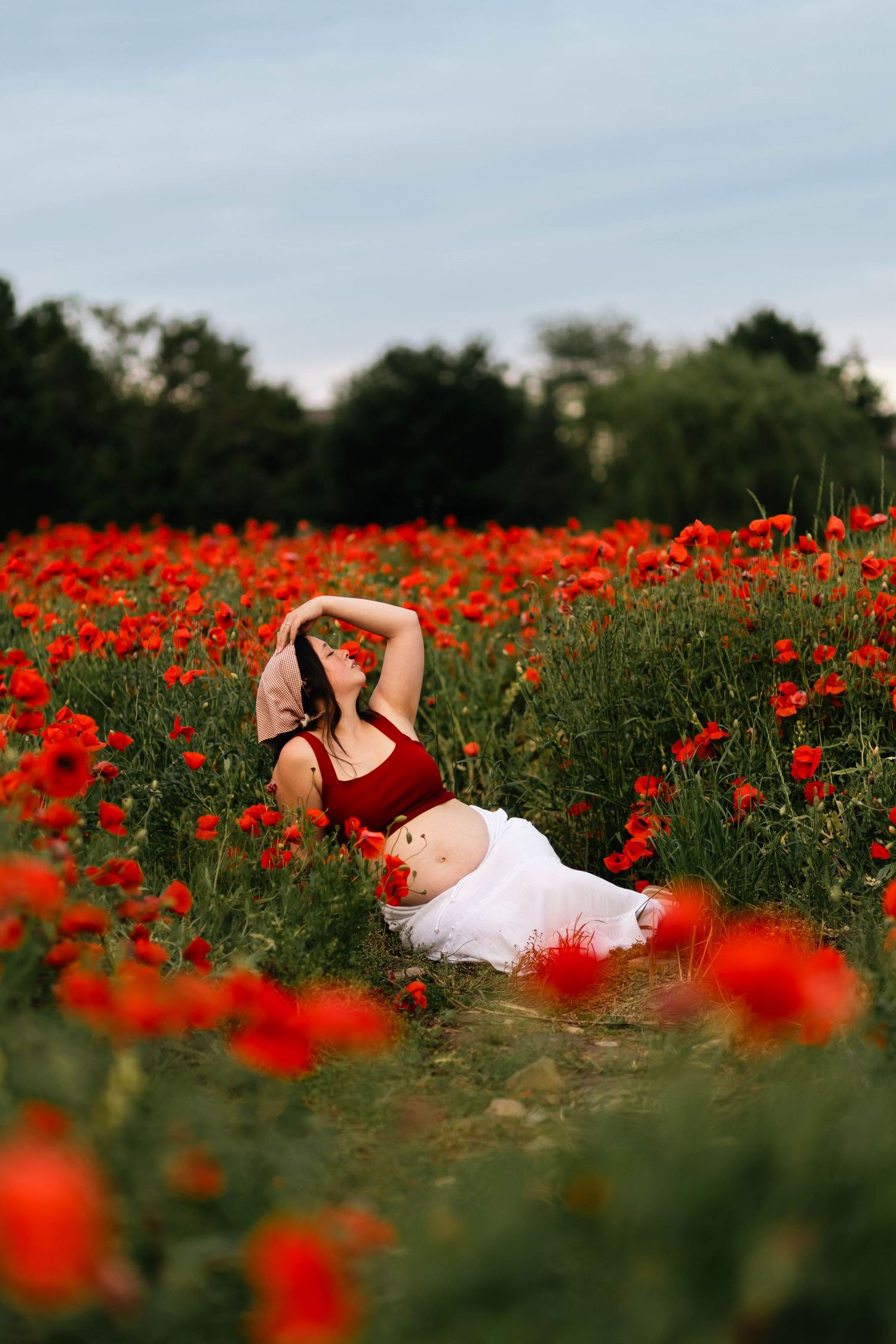 A pregnant woman in a red crop top and white skirt reclines among vibrant red poppy flowers in a field in Douglassville, PA.