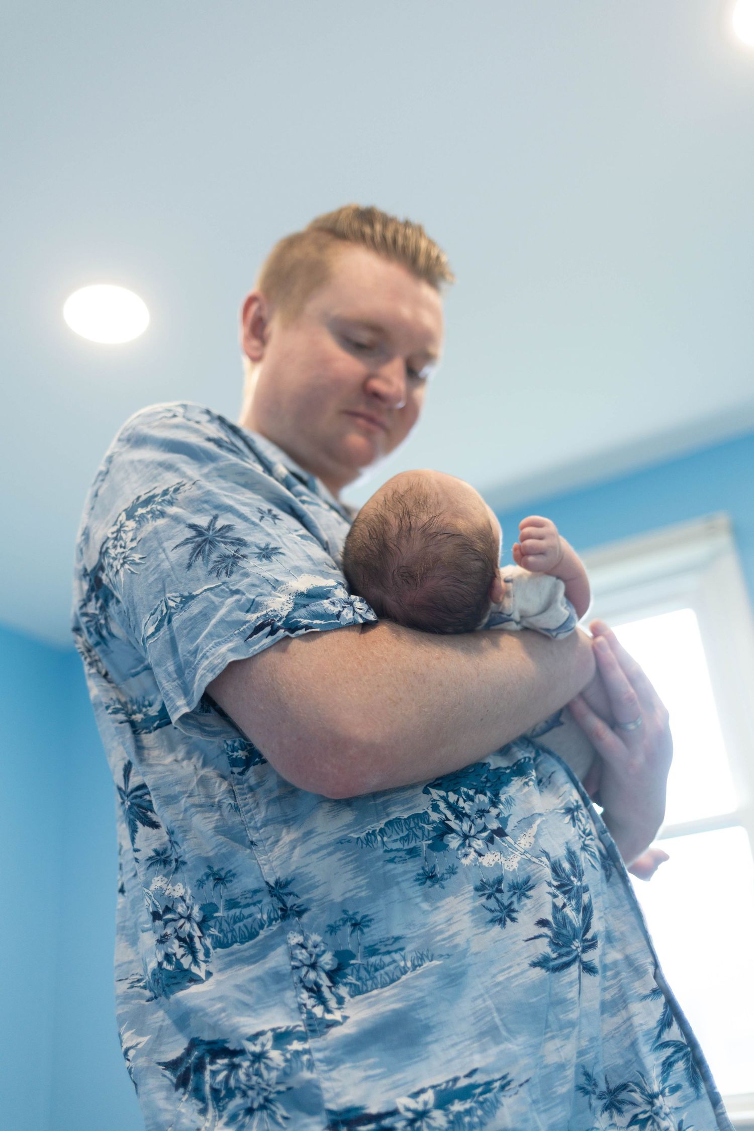 Proud new father in floral shirt cradling sleeping newborn baby against his chest during an at home newborn session Philadelphia, PA.