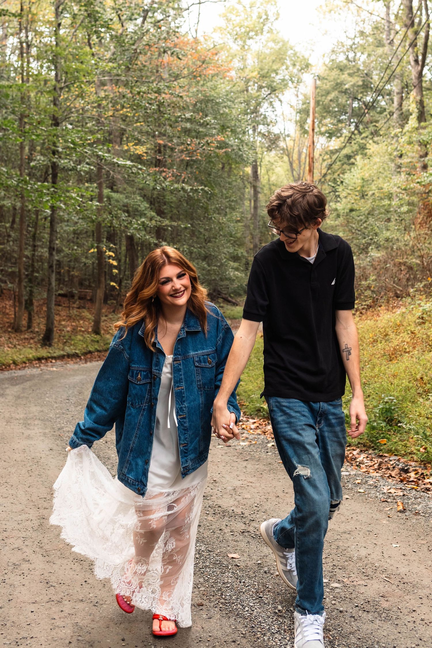 A smiling couple walks hand-in-hand down a forest path, woman in white lace dress and denim jacket, man in black shirt in Oley, PA.