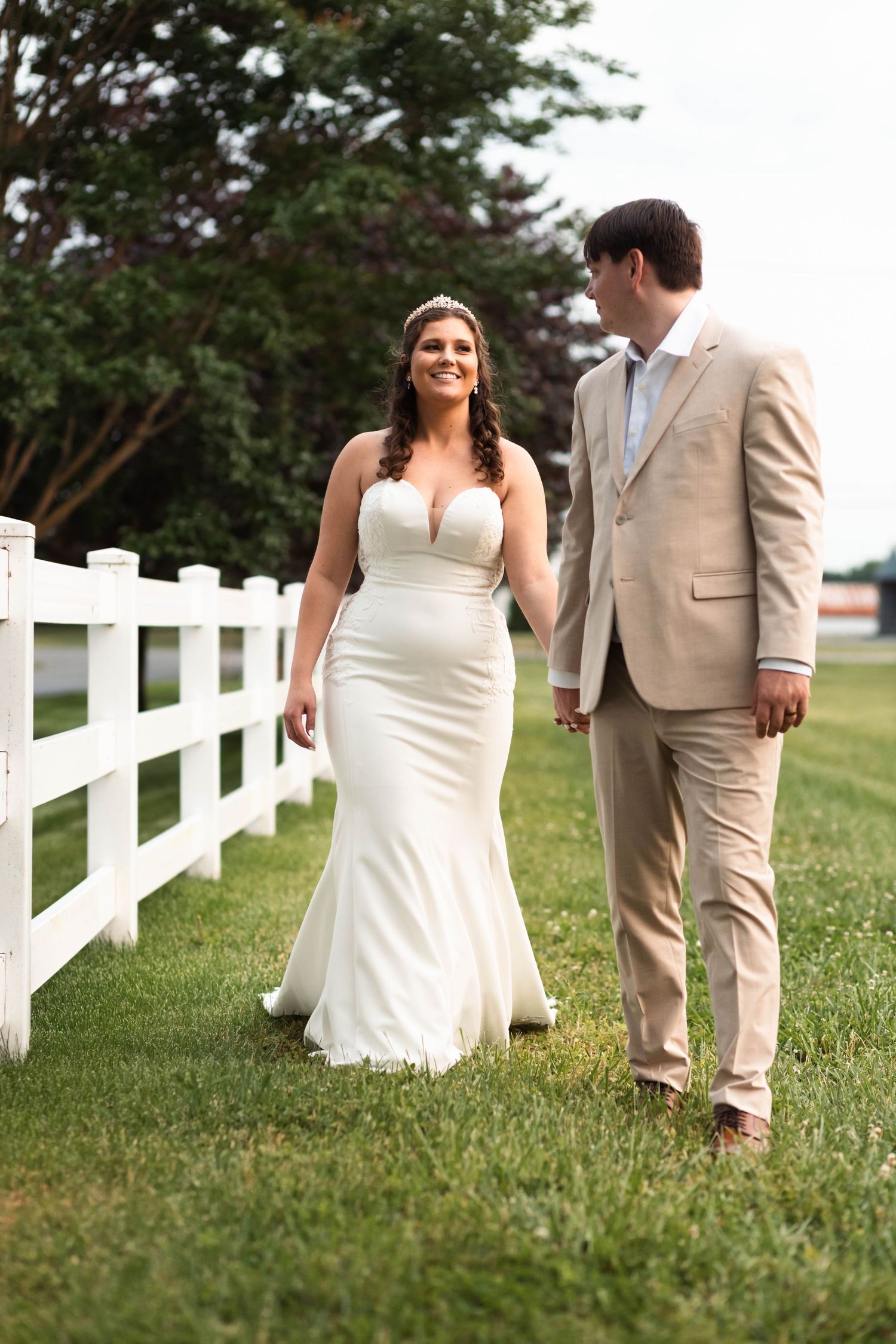 Bride and groom walking hand in hand along a white fence on a green lawn outdoors.