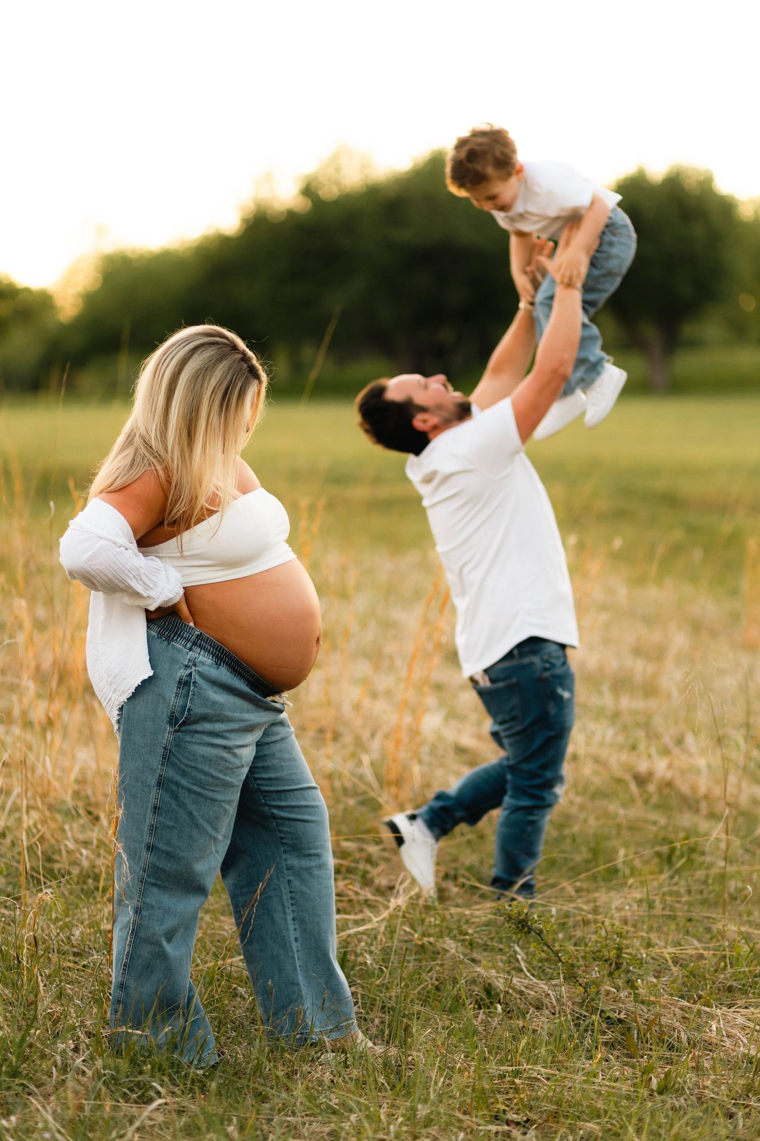 Pregnant woman and husband playing with young son in sunny field during maternity photo session.