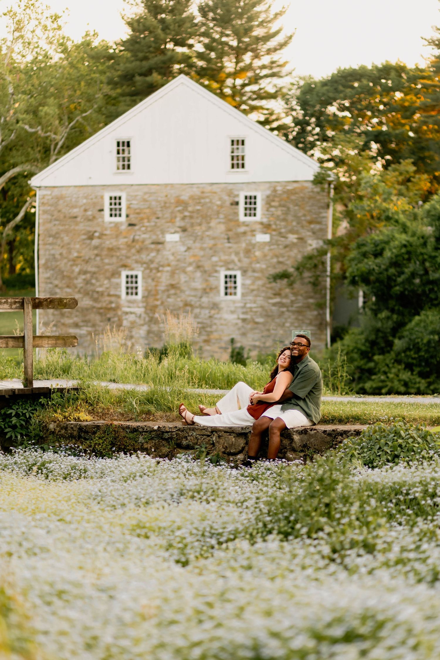 A couple sits together near a pond in front of a historic stone building surrounded by wildflowers at Grings Mill in Reading, PA.