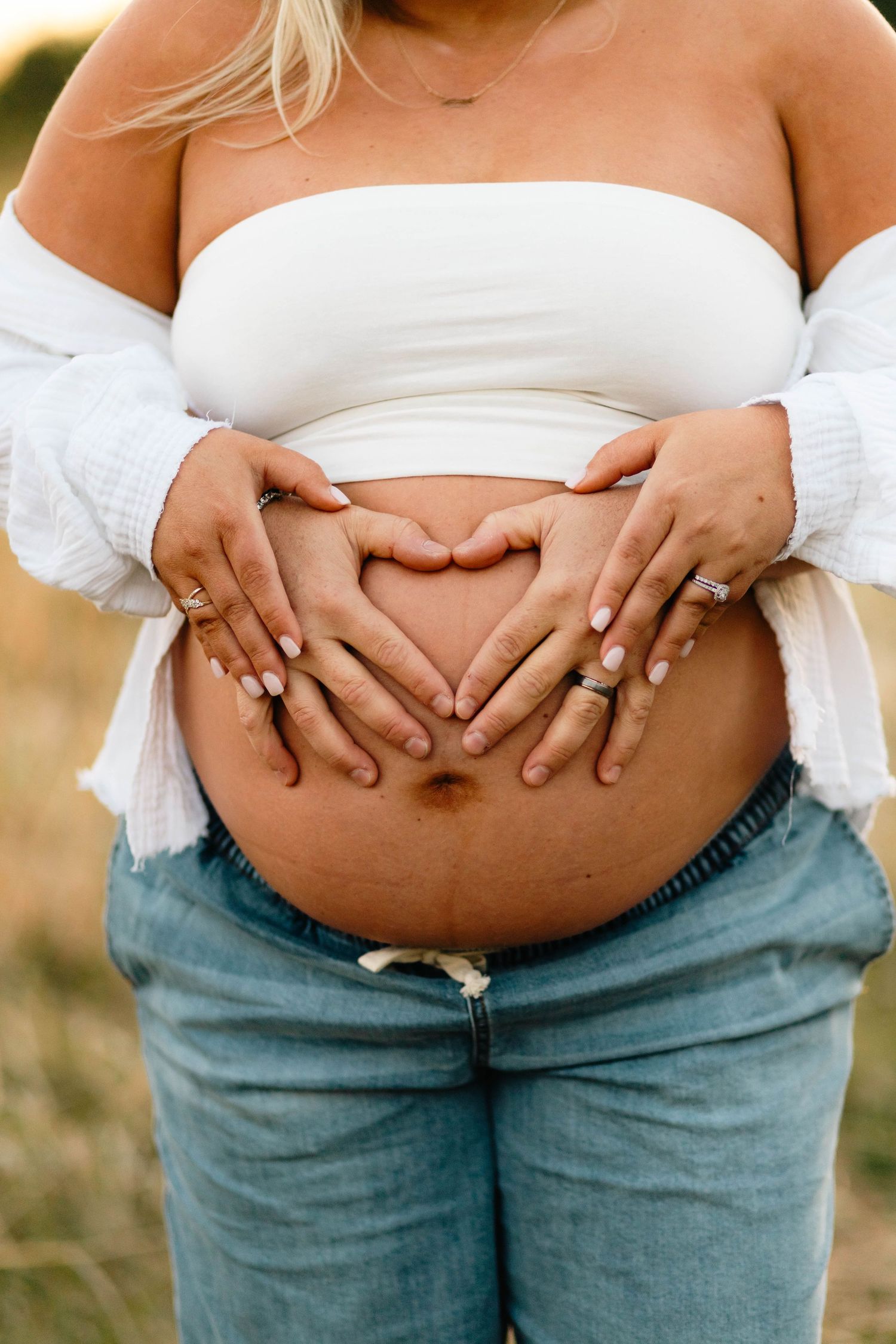 Two pairs of hands forming a heart shape on a pregnant womans bare belly outdoors in natural light at Hopewell Furnace in Elverson, PA.