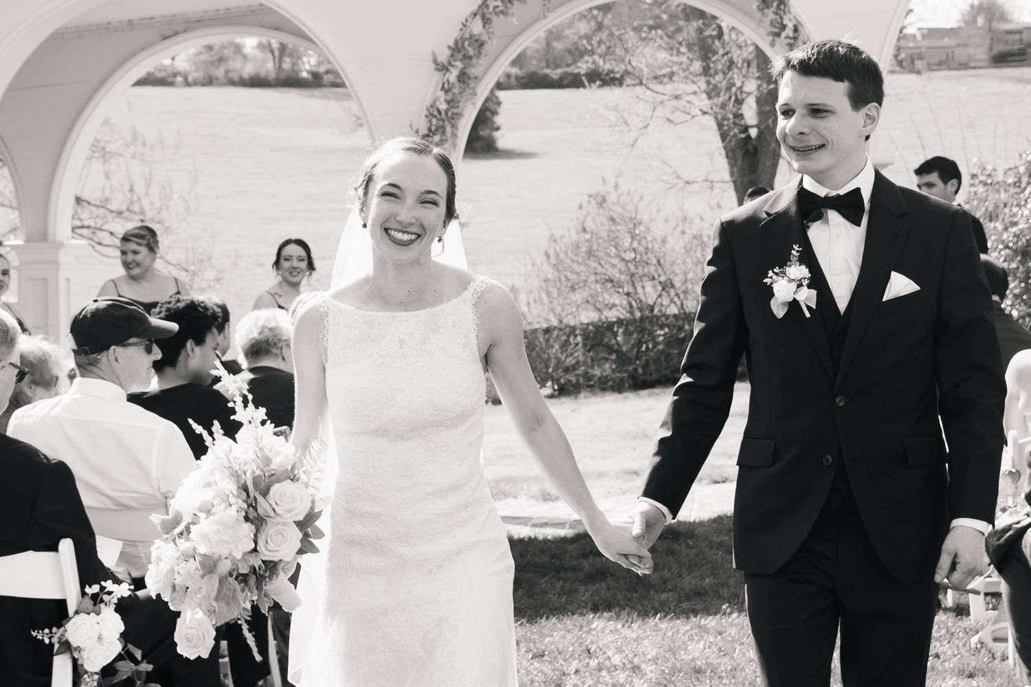 Joyful newlyweds walk hand in hand down the aisle after outdoor wedding ceremony in black and white.