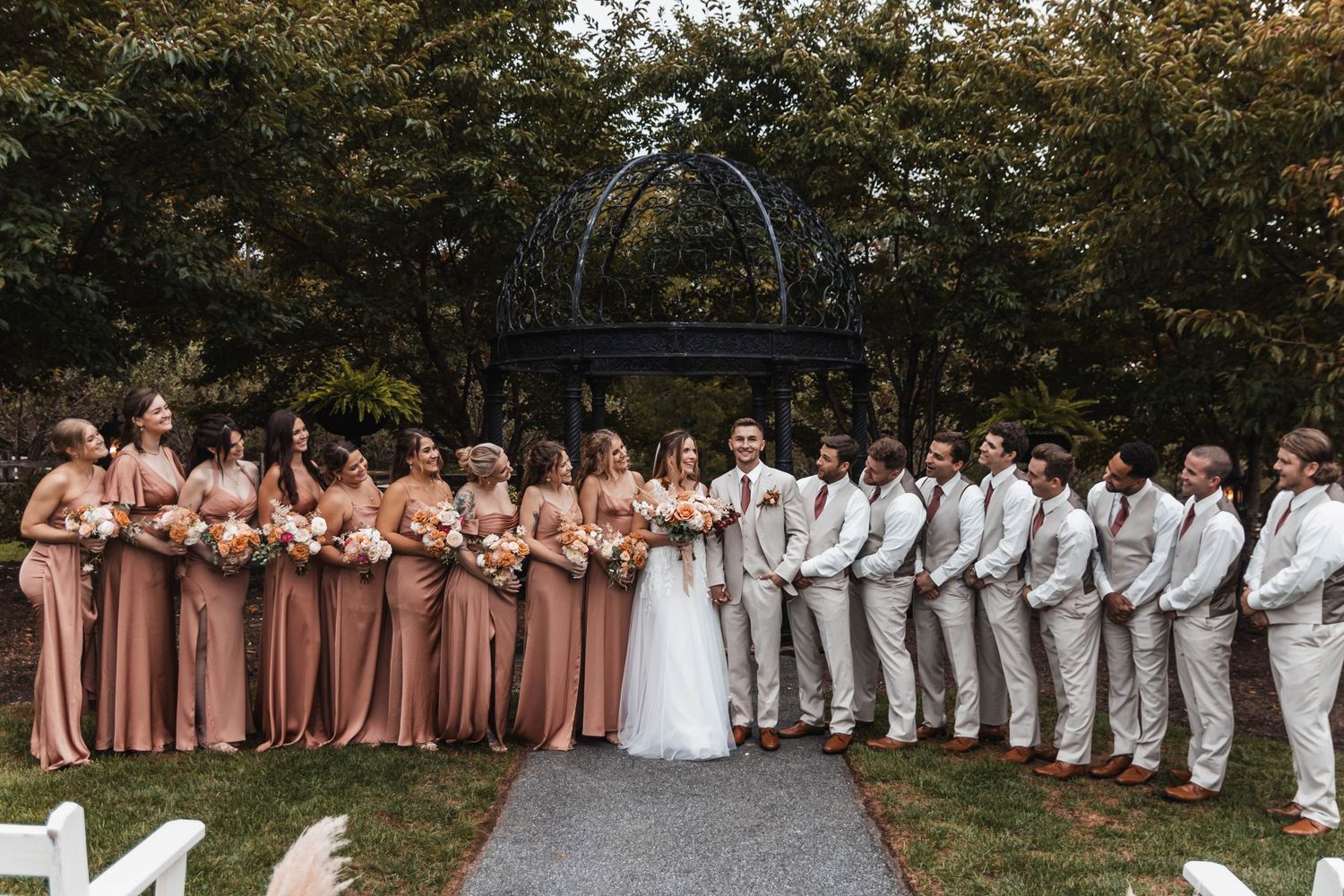 Wedding party lines up outdoors with bridesmaids in dusty rose gowns and groomsmen in light gray suits.