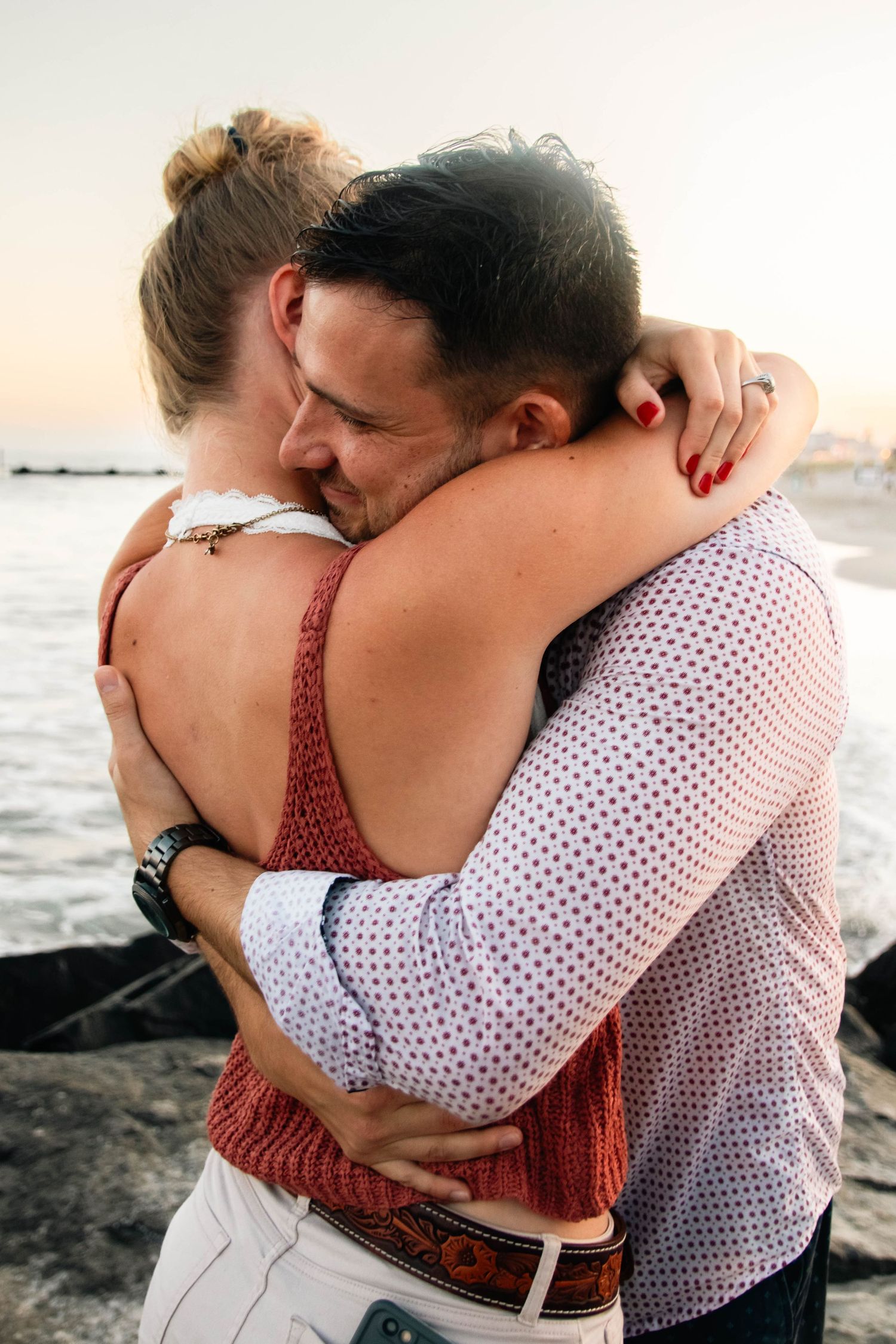 Newly engaged couple sharing a heartfelt embrace on rocky beach at sunset after a marriage proposal.