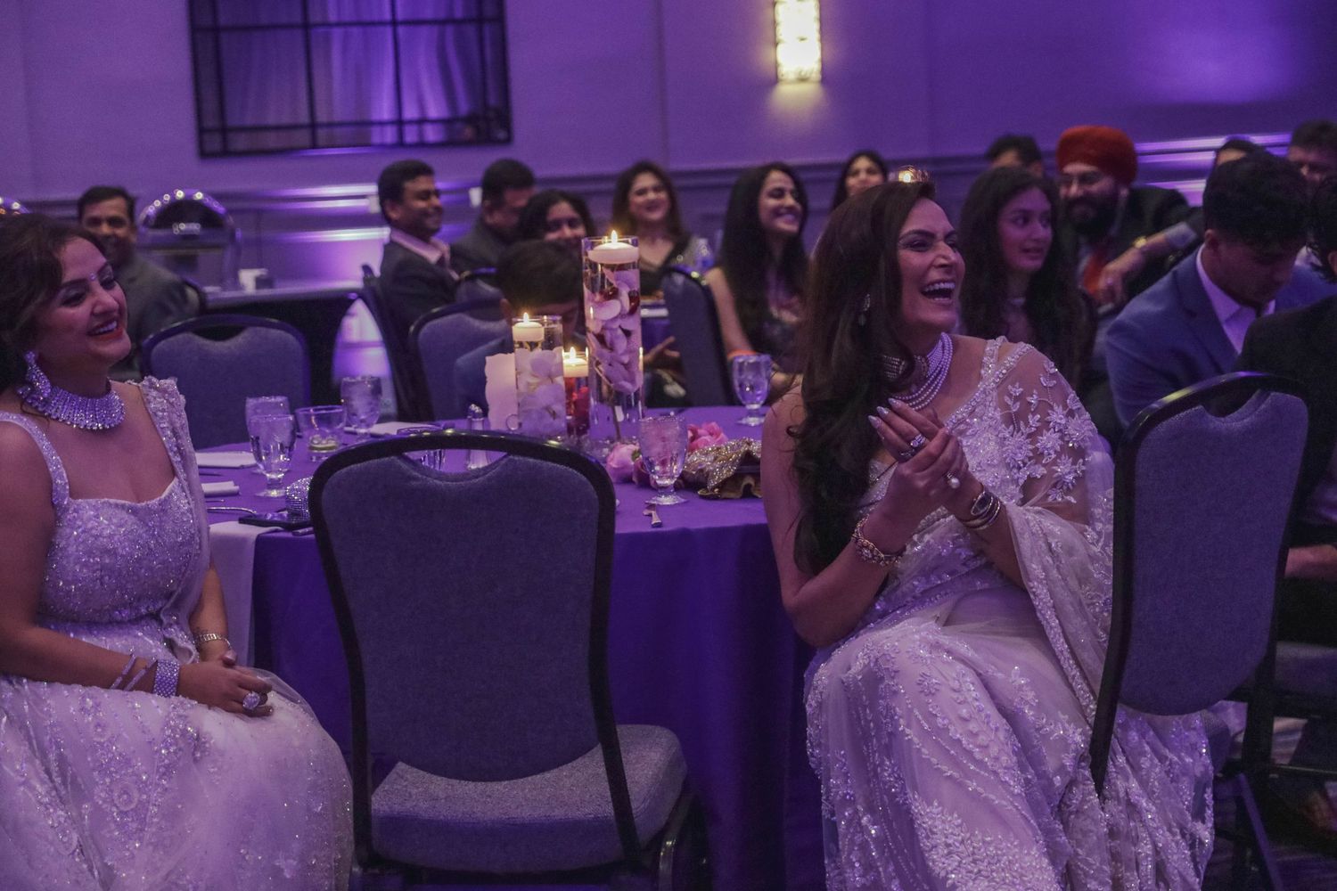 Guests in white and silver sarees laughing at tables during an elegant Indian wedding reception.