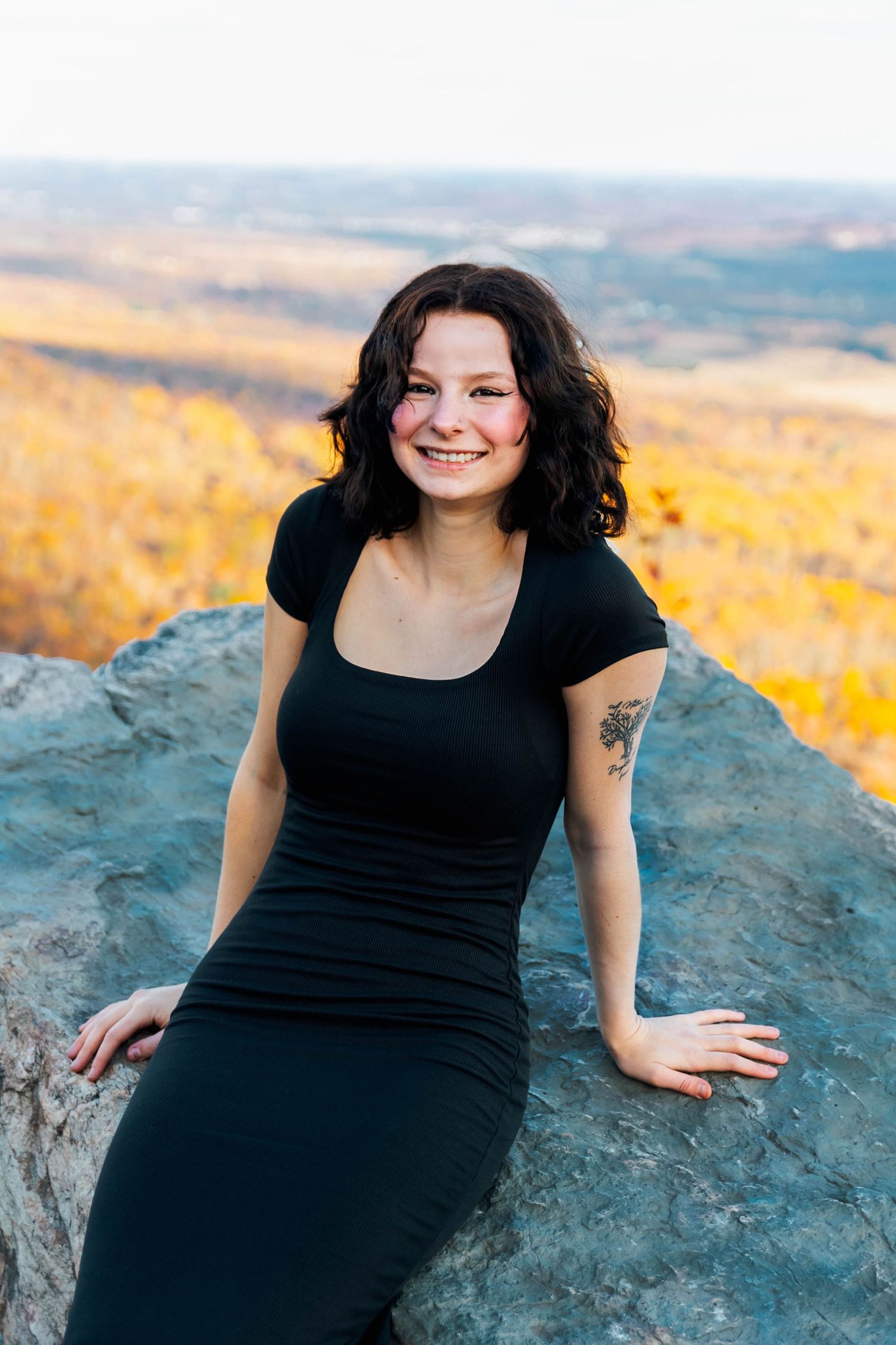 Smiling brunette woman in black dress posing on mountain rock with autumn valley view.