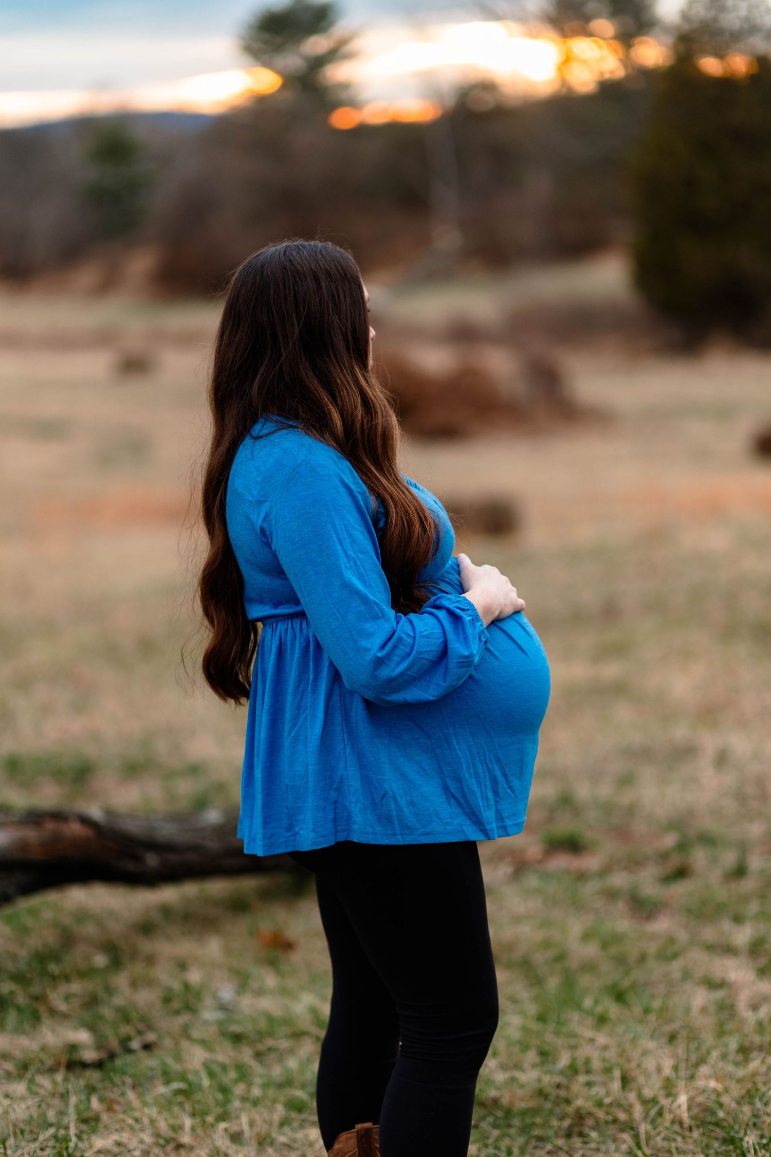 Pregnant woman blue top holding baby bump while standing in field during sunset maternity shoot at Daniel Boone Homestead.