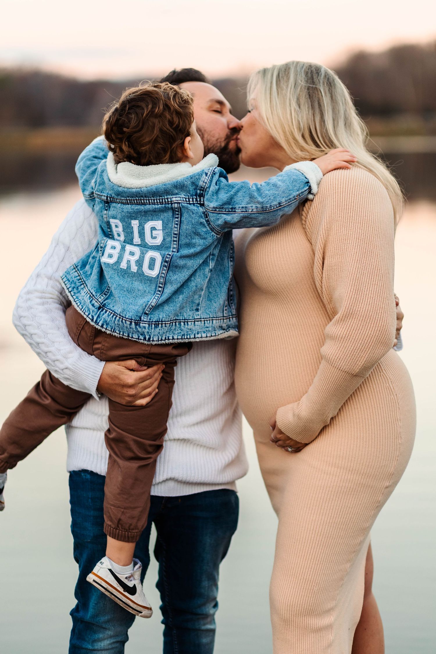 Pregnant woman in beige dress kissing partner holding toddler in Big Bro jacket at Green Lane Park.