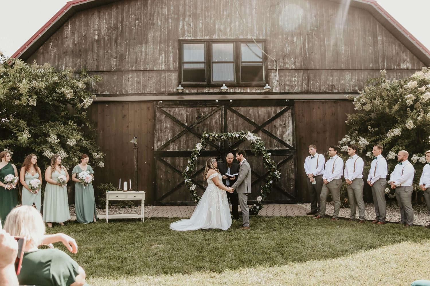 Wedding ceremony in front of a rustic barn with bridesmaids in sage green and groomsmen in gray suits.