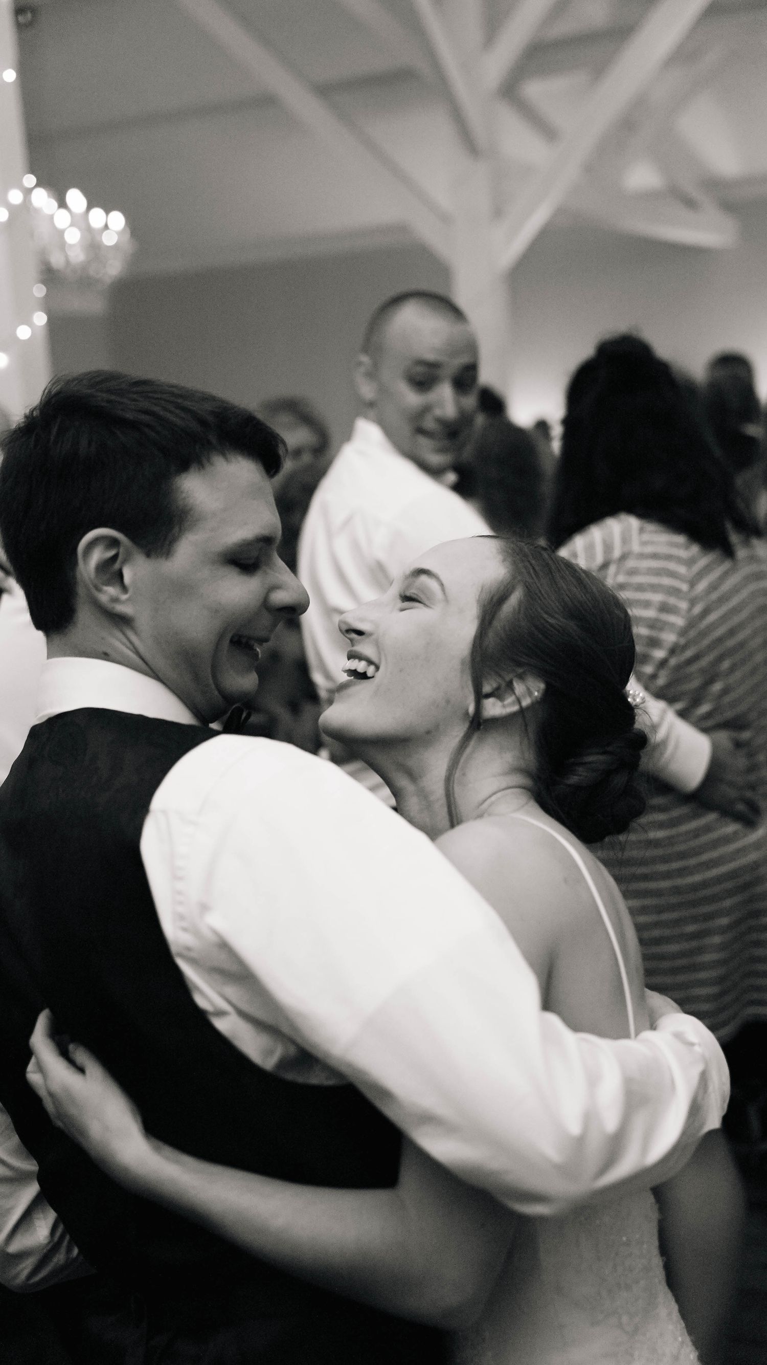 Black and white photo of laughing bride and groom embracing joyfully on crowded wedding reception dance floor.