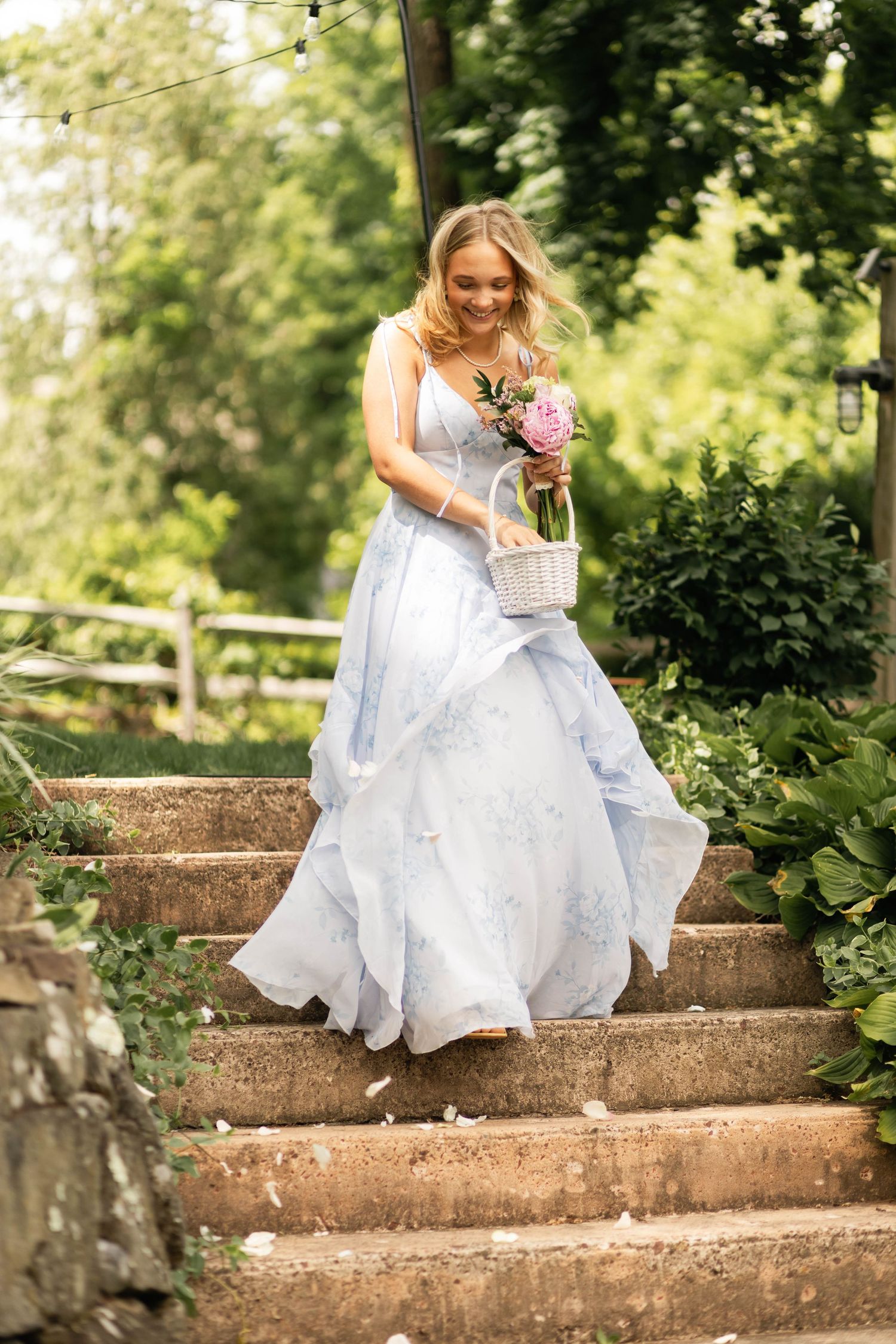 Smiling bride in a light blue floral gown carrying a flower basket while descending stone steps outdoors.