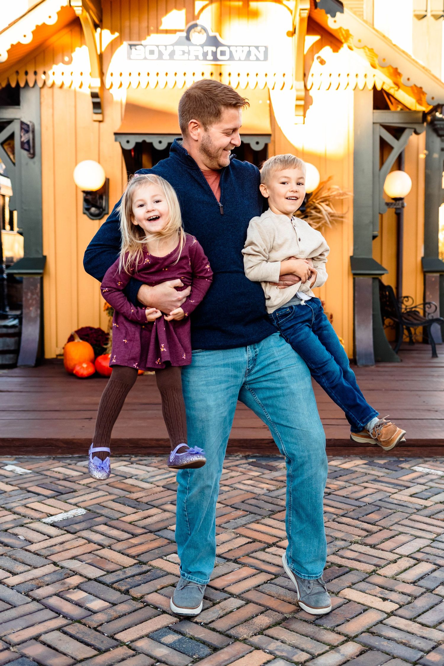 A cheerful father lifts his two young children playfully outside a warmly lit building at dusk at the Colebrookdale Railroad Station.