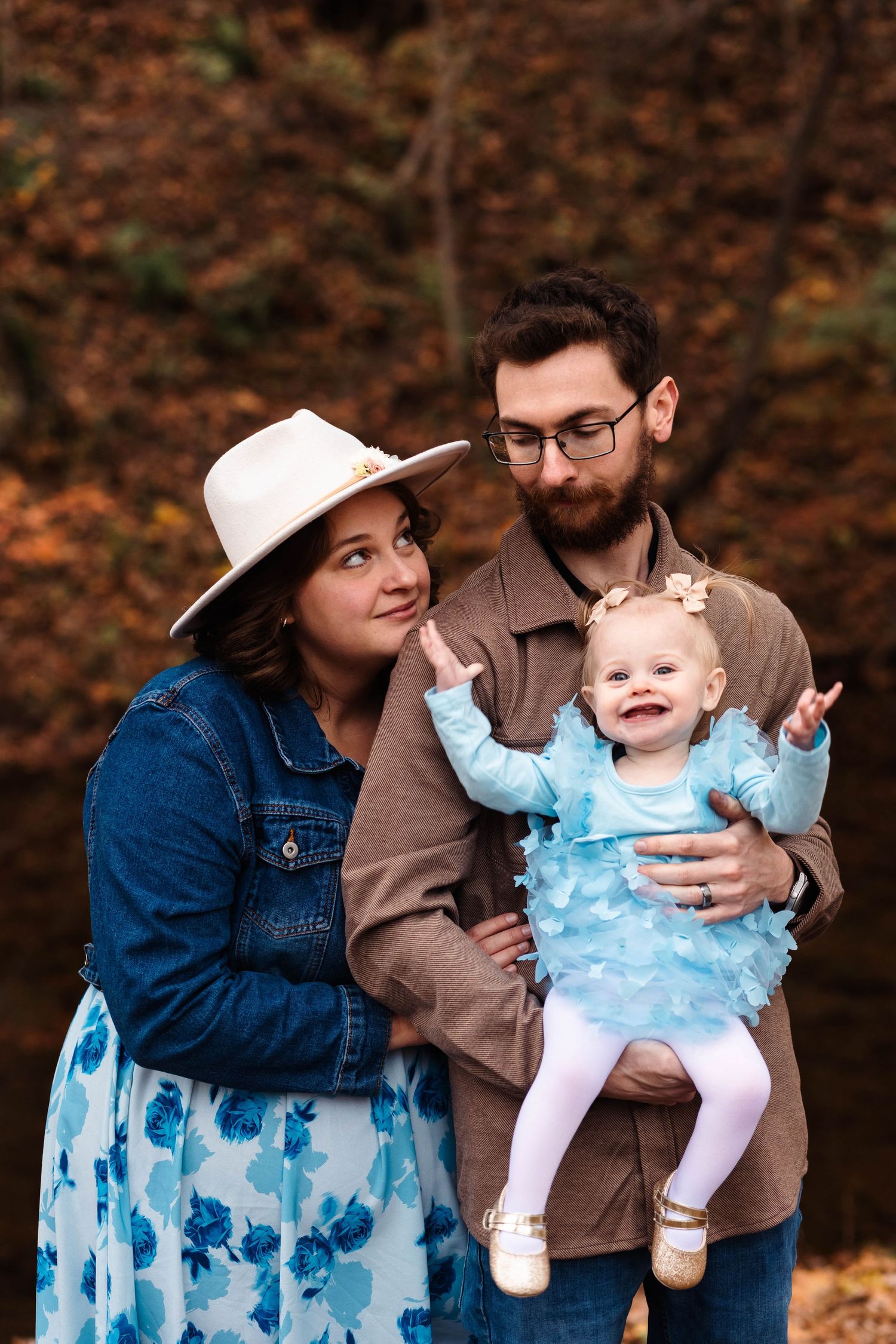 Bearded father holds smiling baby in blue dress while mother in hat and denim jacket poses outdoors in Boyertown, PA.
