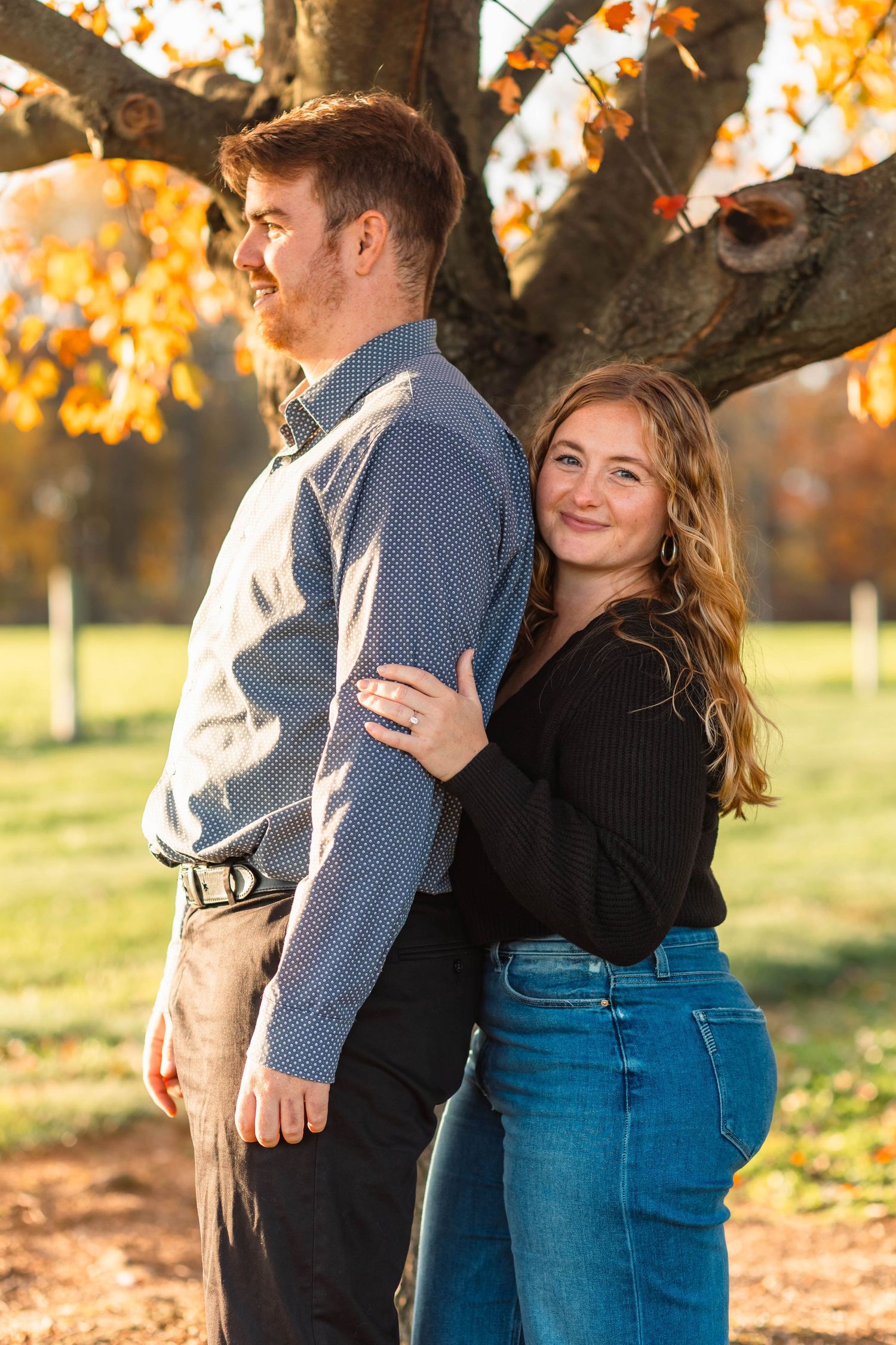 Engaged couple posing back to back under a large autumn tree with golden fall foliage backdrop in Lancaster, PA.