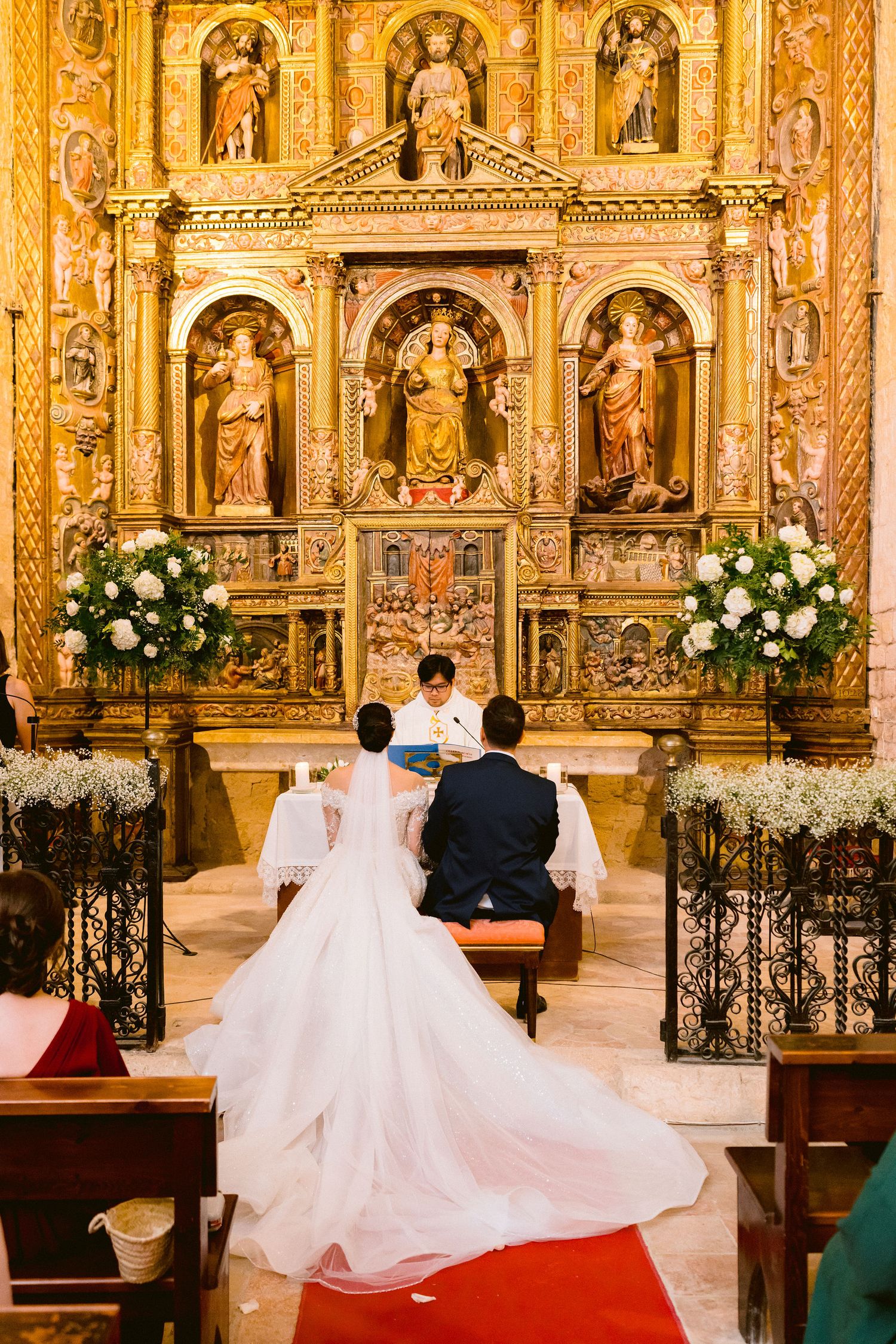 Traditional church wedding ceremony taking place in an ornate cathedral with gilded altars and white floral decorations.