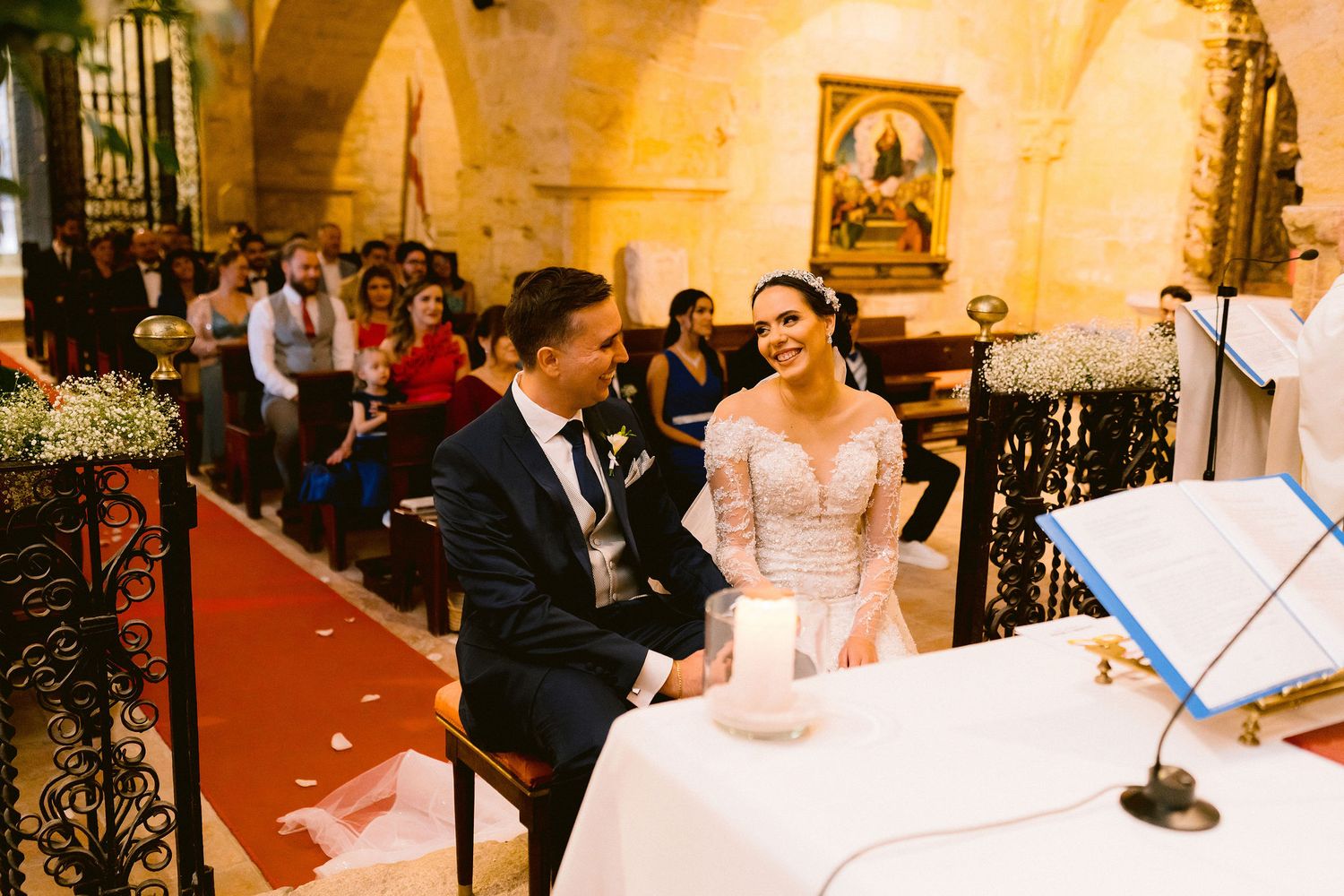 Wedding couple sits at an ornate ceremony table inside a warmly lit historic chapel with religious artwork and architecture.