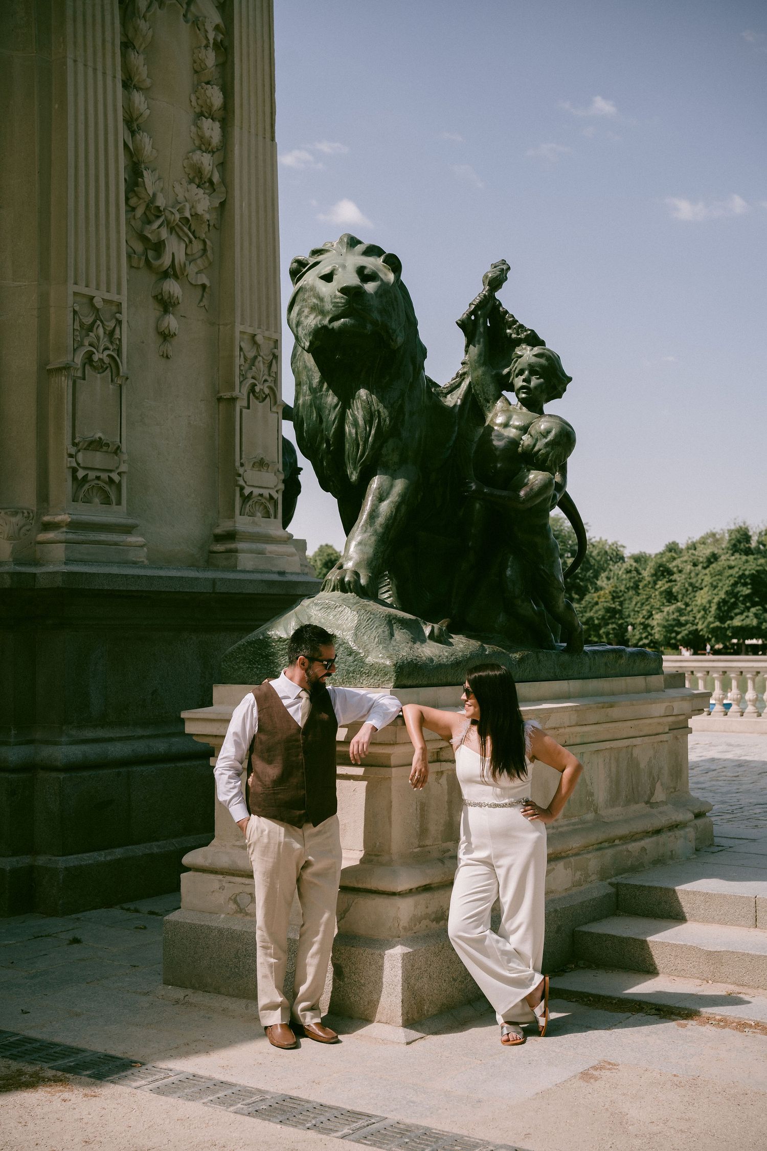 A couple poses by a decorative lion statue near classical architecture.