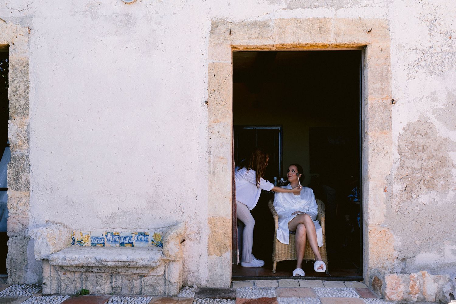Two figures sit in a rustic doorway of an old stone building with weathered white walls.