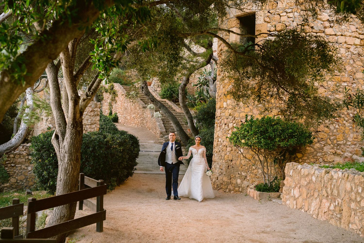 Elegant couples take formal wedding photos along a stone pathway surrounded by lush greenery.