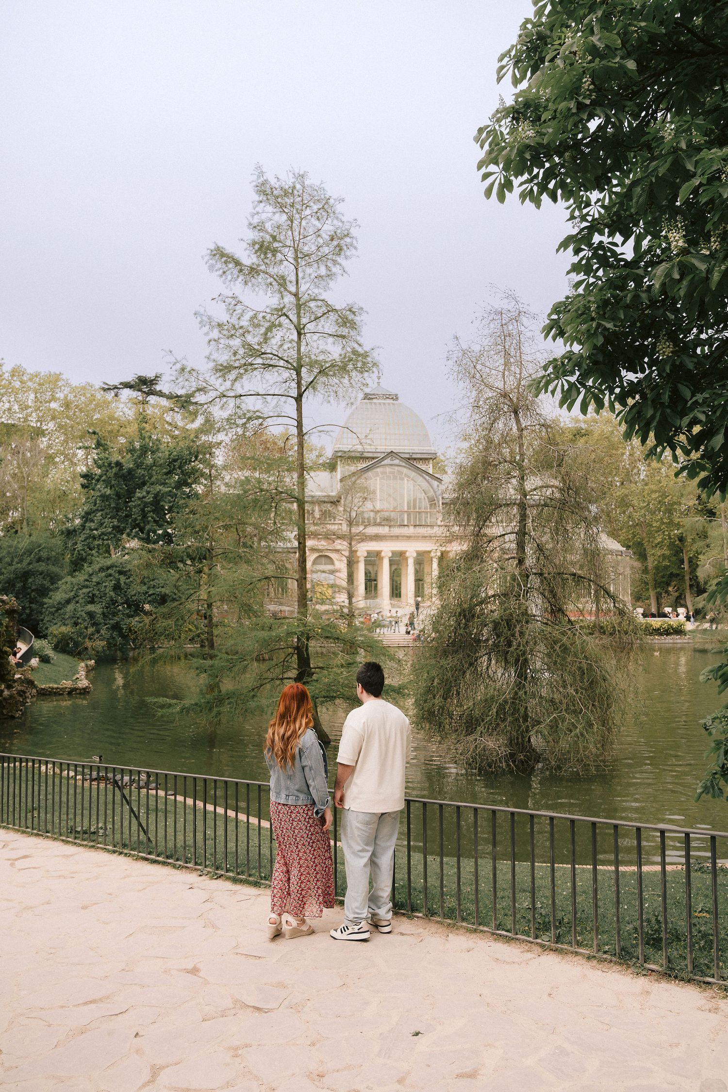 A couple admires the historic architecture of Retiro Park Palace and lake in Madrid on an overcast day.