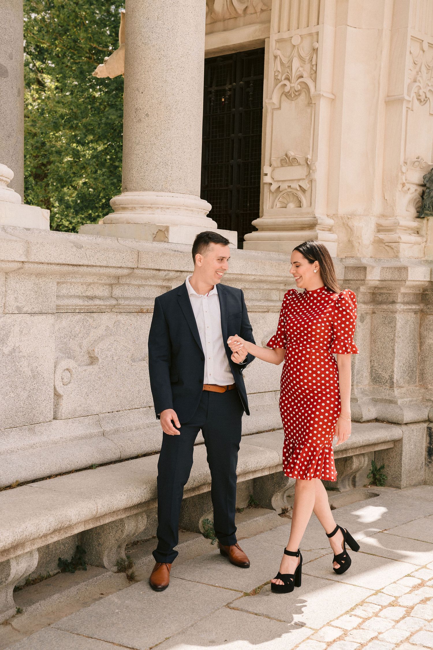 A couple poses playfully along a classical stone colonnade with ornate architectural details.