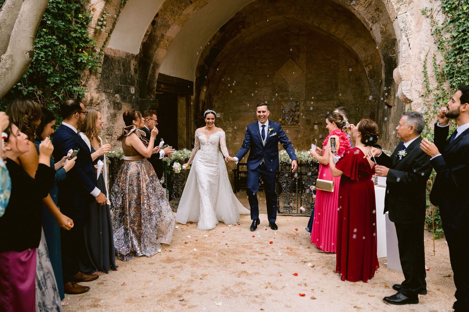 Wedding celebration with guests lined up along stone archway path throwing flower petals during joyous ceremony exit.