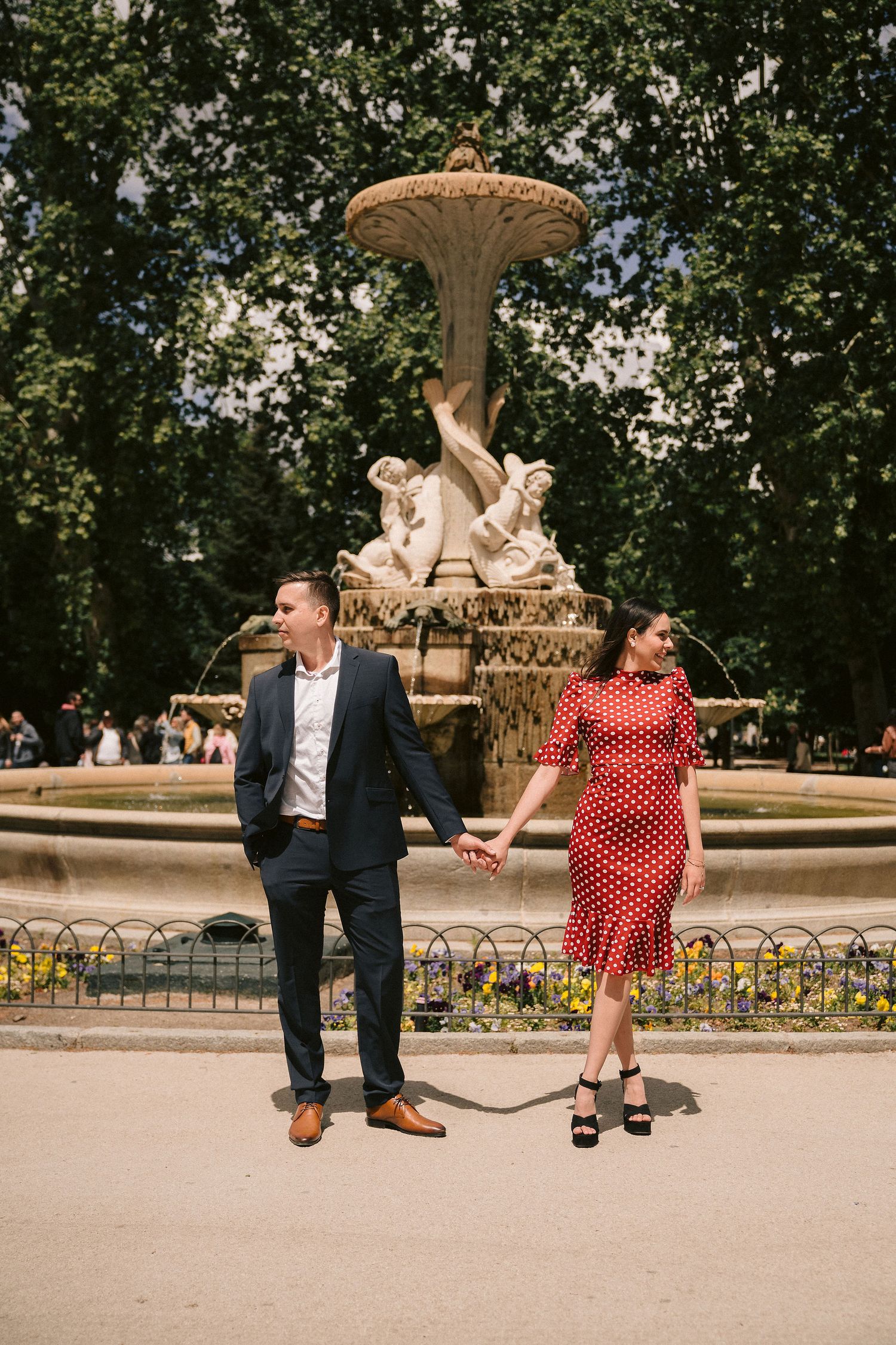 A couple sits and poses by an ornate fountain in a formal garden setting.