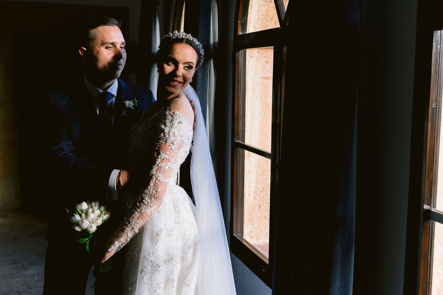 A couple in wedding attire stand together by large windows in a darkly lit indoor setting.