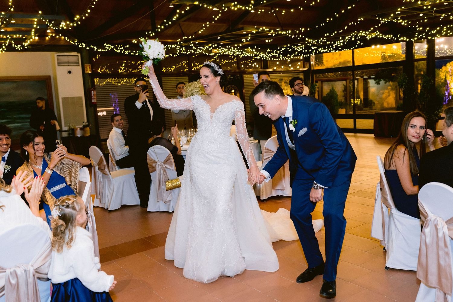 Wedding guests dance and celebrate together in an indoor venue decorated with twinkling string lights overhead.