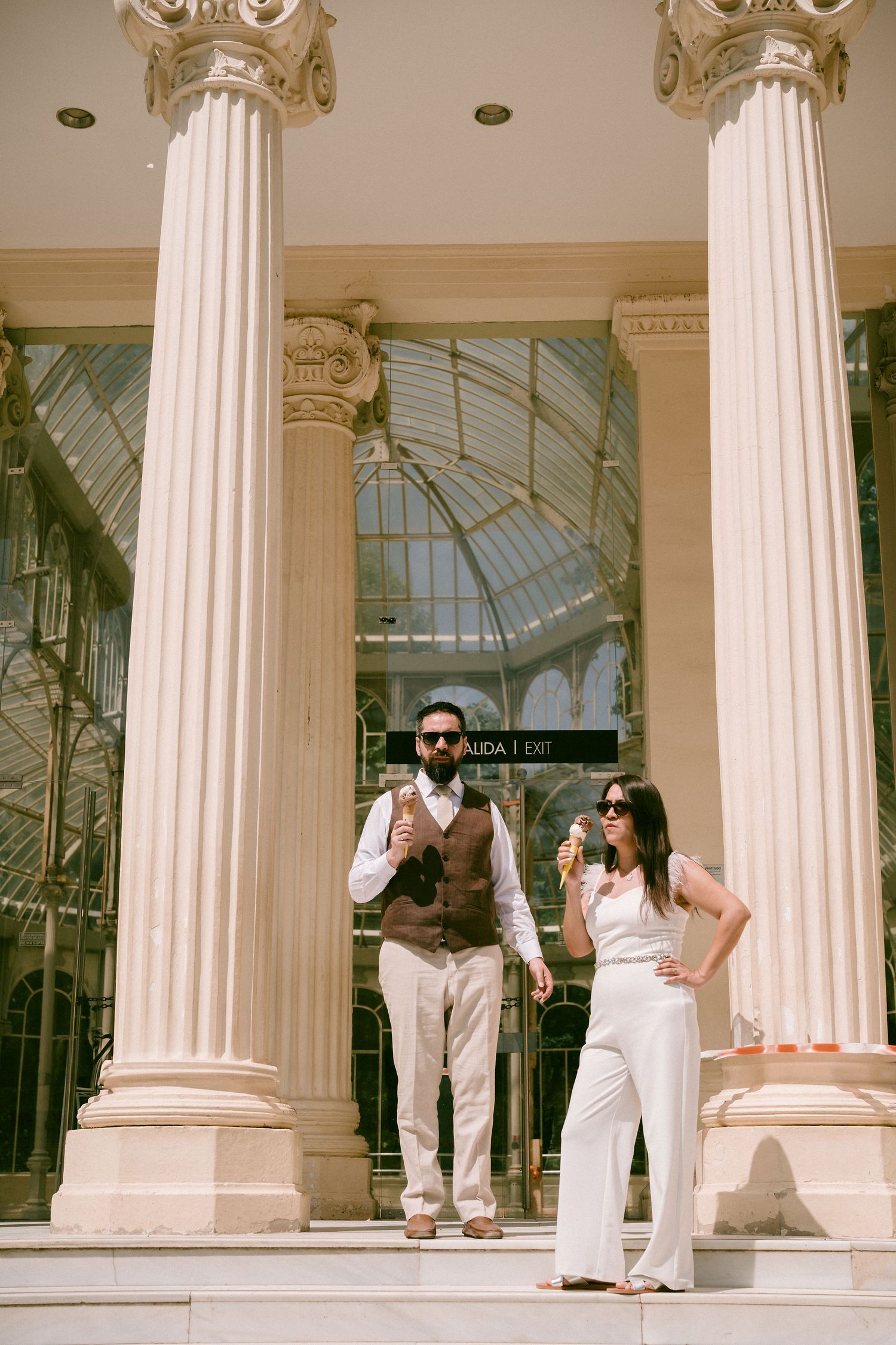 A couple poses between ornate white columns of a glass-roofed building.