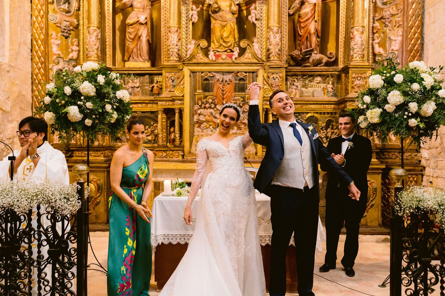 Wedding ceremony in ornate church with floral arrangements featuring white roses and golden altar decorations.