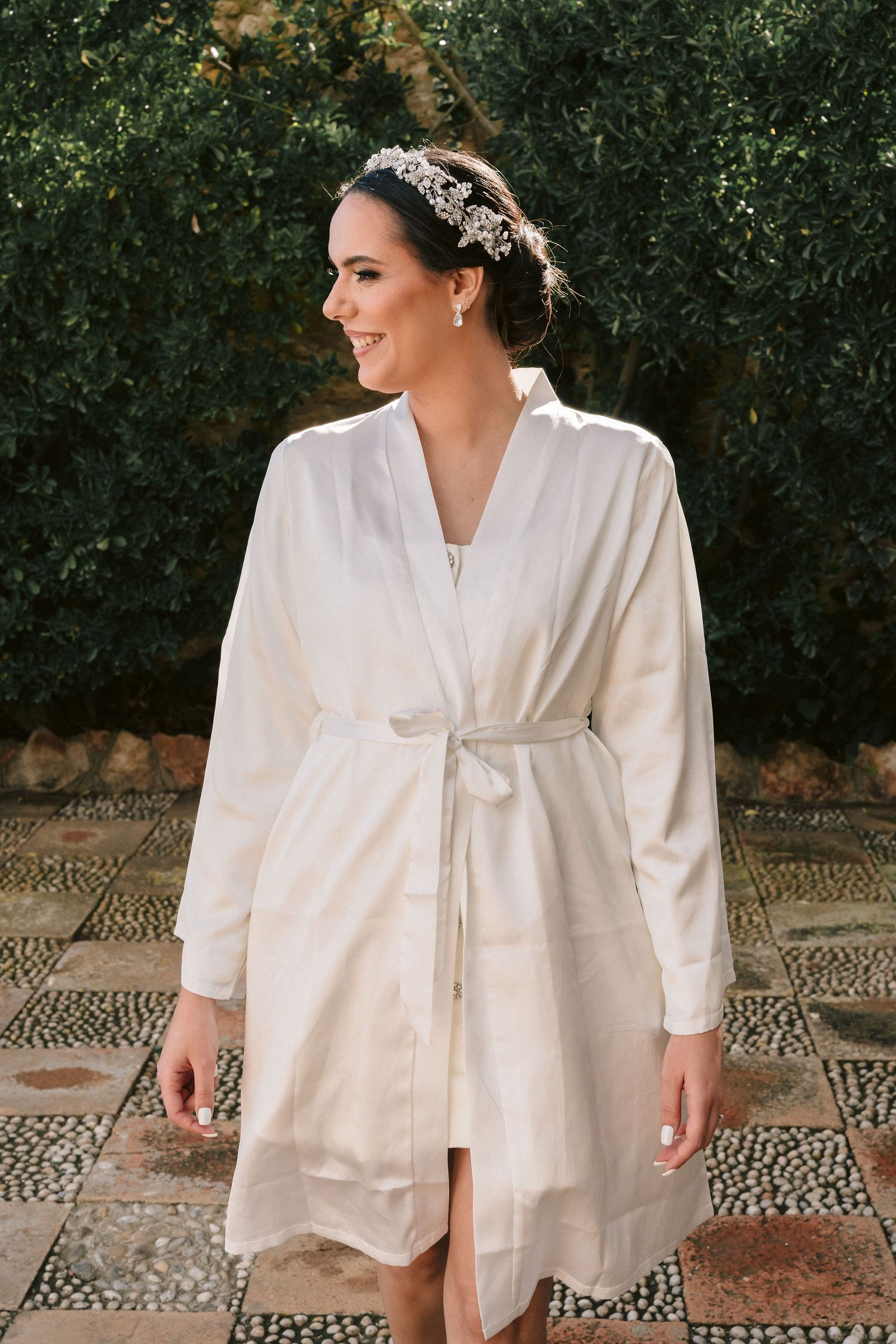 A person wearing an elegant white flowing robe with a matching sash and ornate headpiece poses in a garden setting.