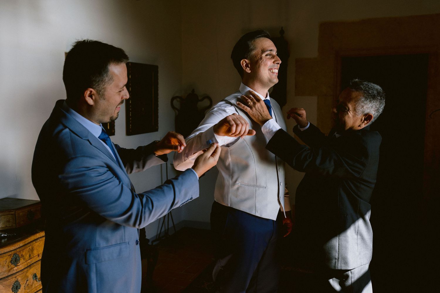 People help adjust a light blue tie while getting ready in a dimly lit room before a formal event.