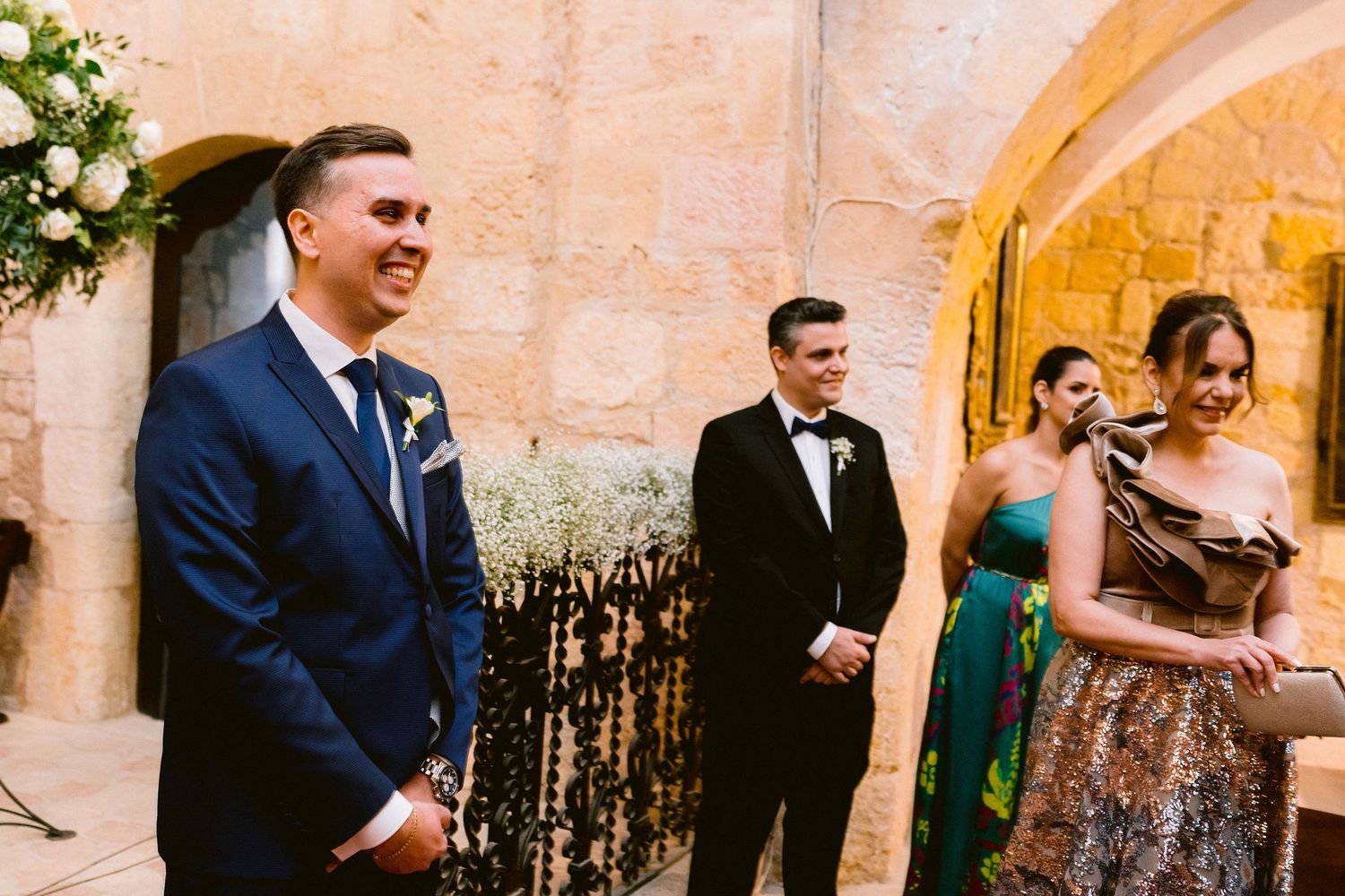 Wedding party members in formal attire smile during a ceremony at an elegant stone venue with warm lighting.