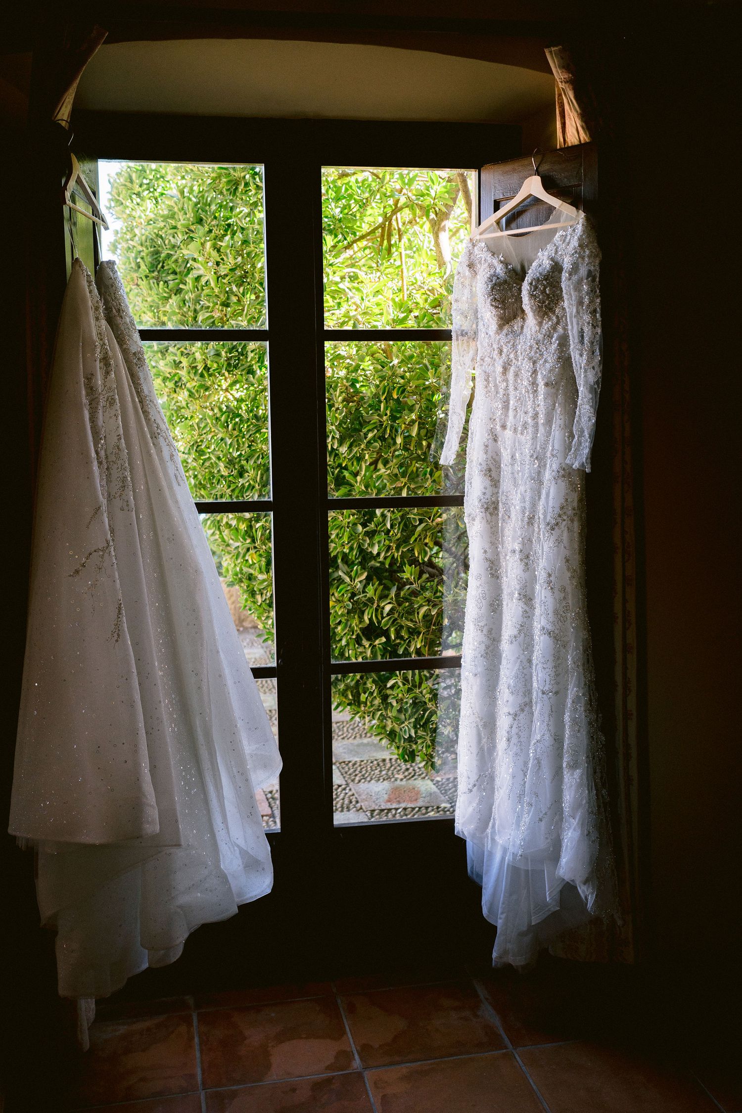 Two wedding dresses hang elegantly by a sunlit window frame, one lace gown and one traditional white dress.