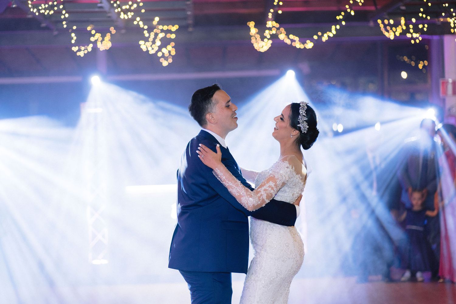 Newlyweds share their first dance at an elegant wedding reception with romantic lighting and twinkling string lights overhead.