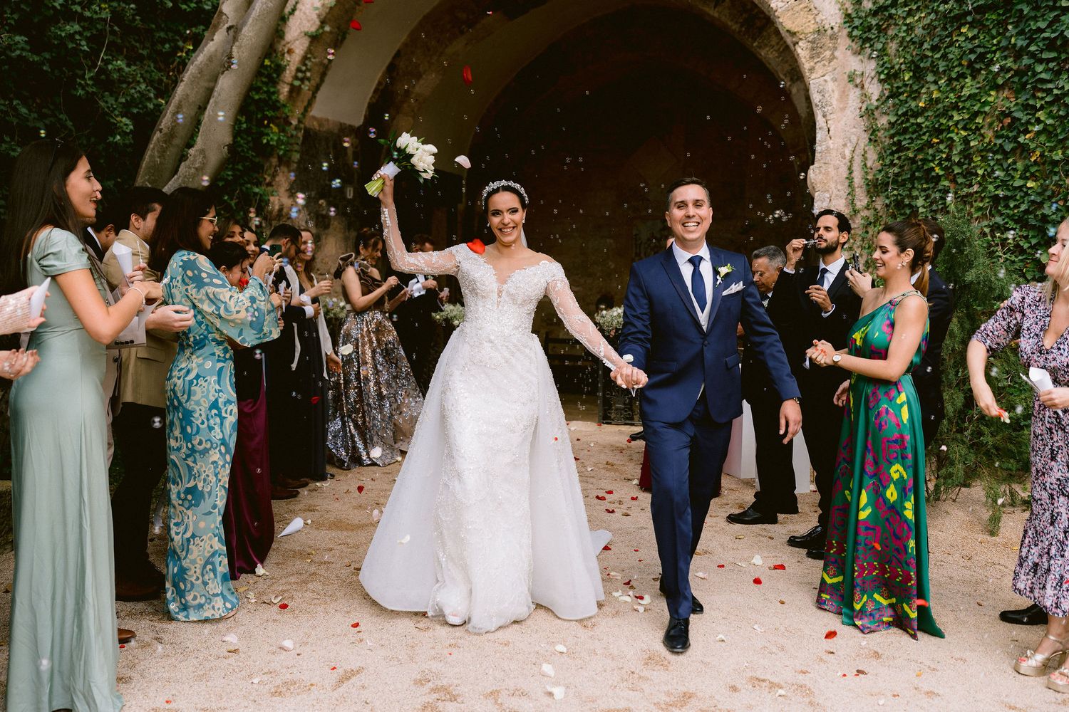 A newlywed couple exits through a stone archway as wedding guests throw white confetti and celebrate their union.