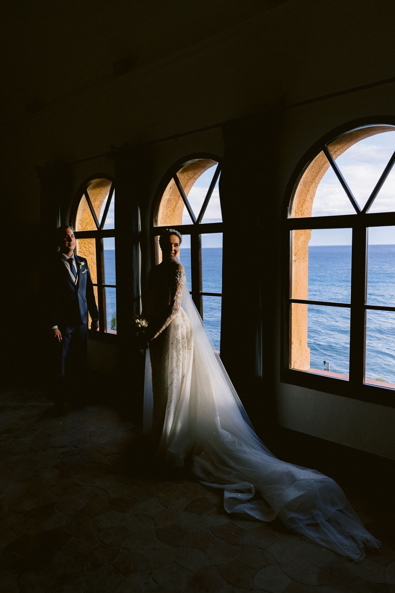 A flowing white wedding dress against dark silhouettes and windows with ocean views creates a dramatic scene.