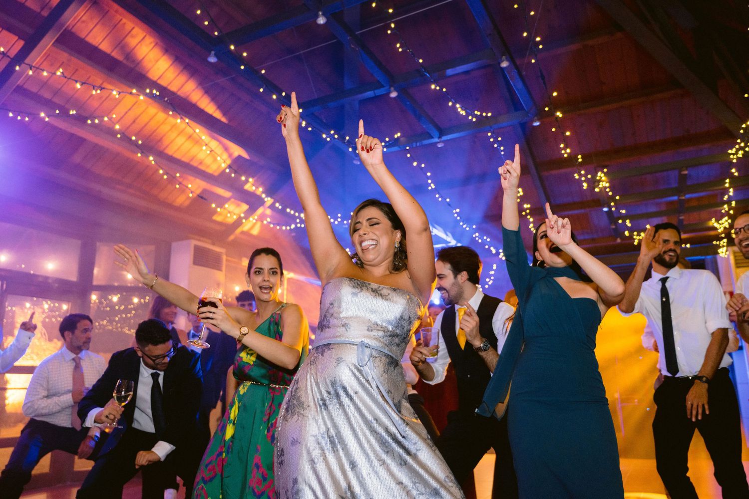 Wedding reception guests dancing and celebrating with raised arms under colorful lighting and string lights in indoor venue.