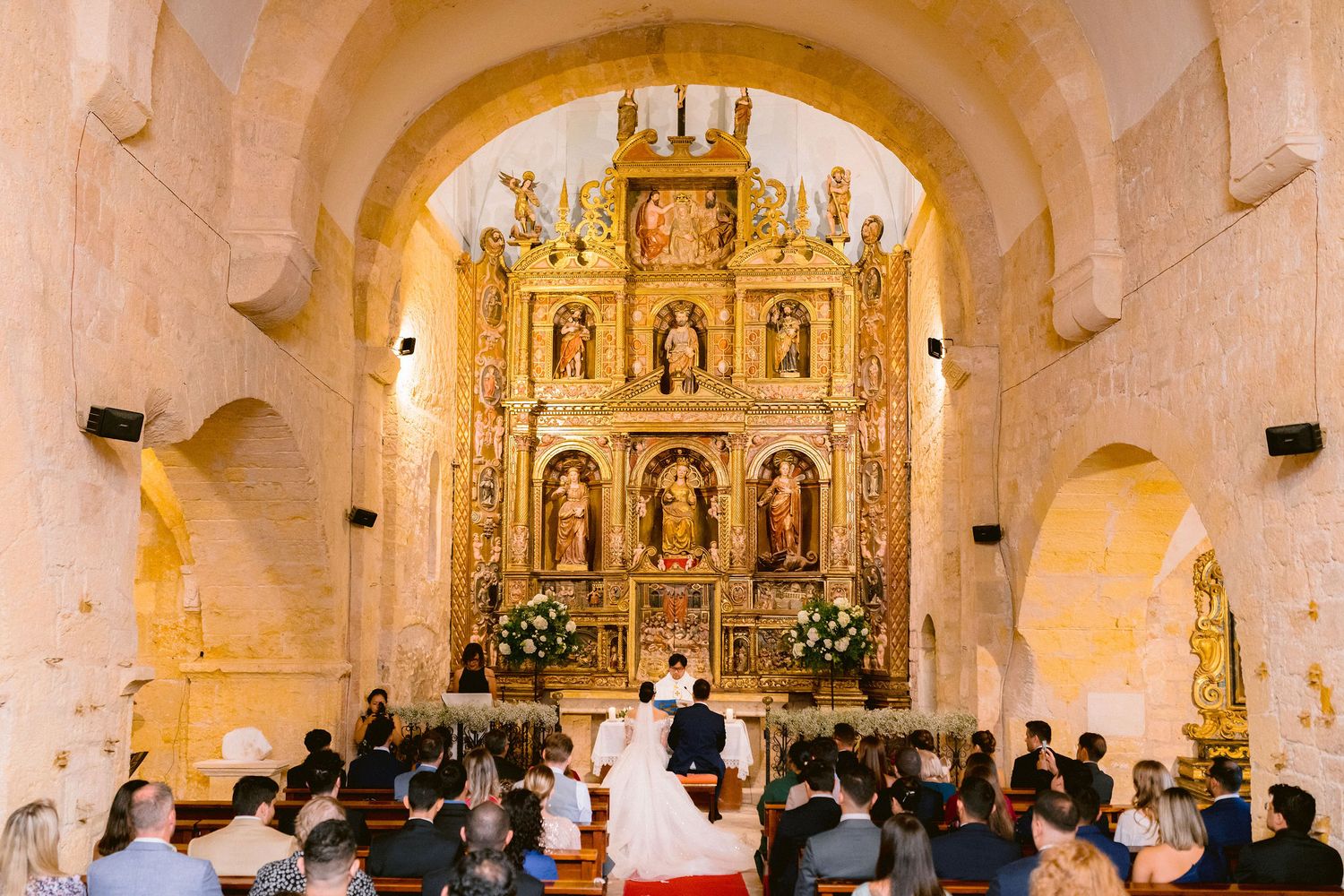 Panoramic view of a Catholic church interior showing ornate golden altar and arched stone walls illuminated in warm light.