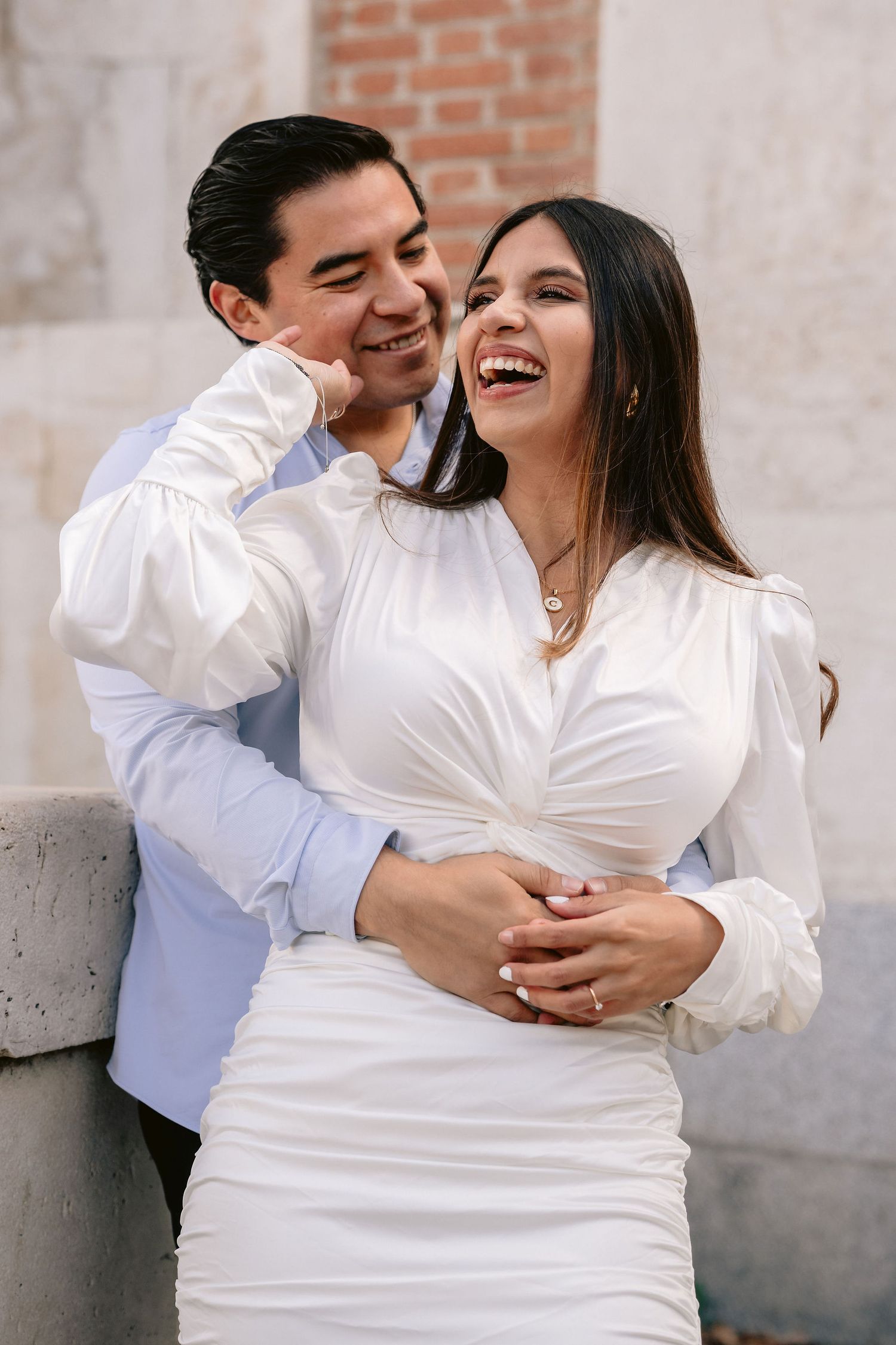 A joyful couple in white outfits share a romantic embrace against a brick wall backdrop.