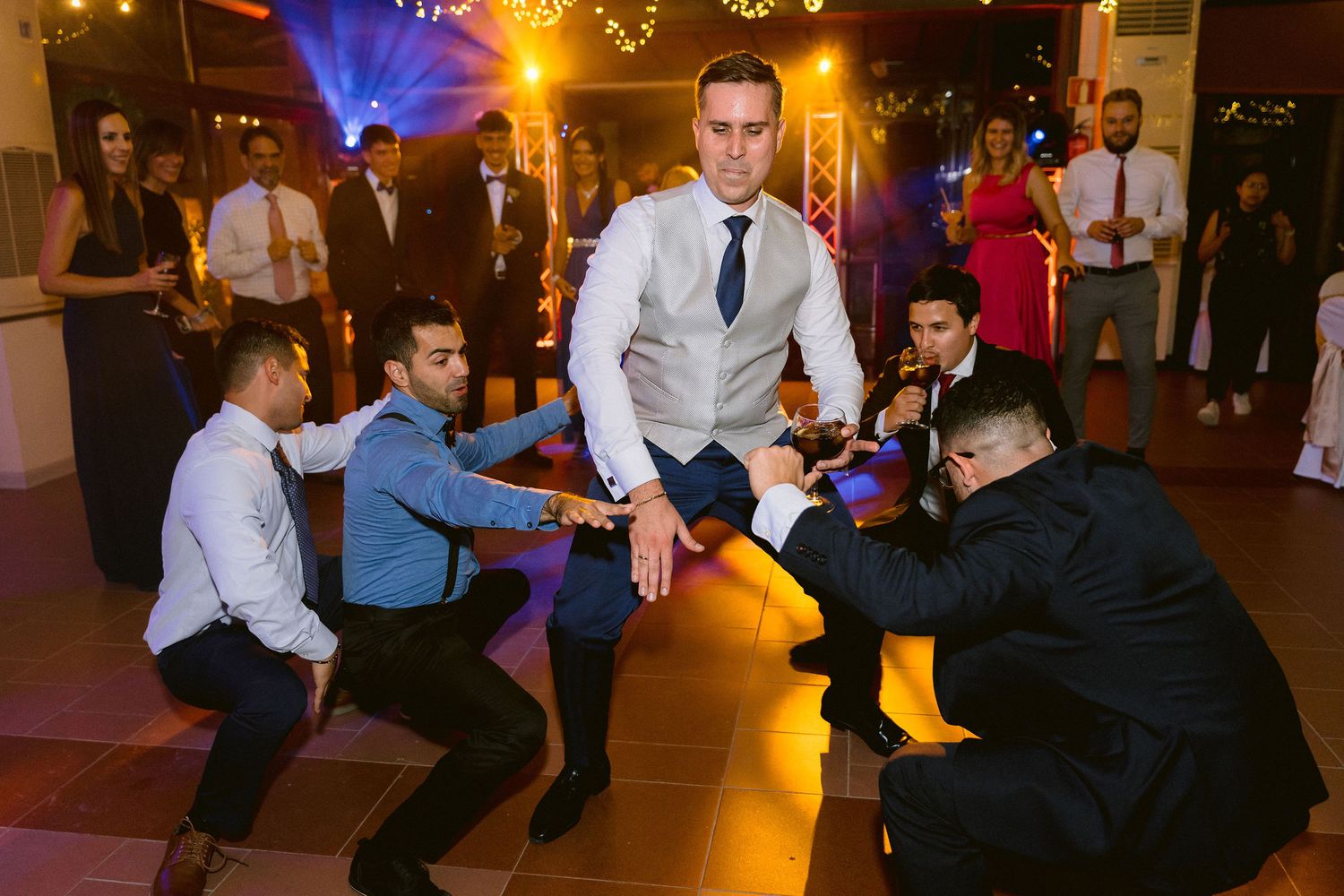 At a lively wedding reception, guests dance in a playful formation with dramatic lighting in the background.