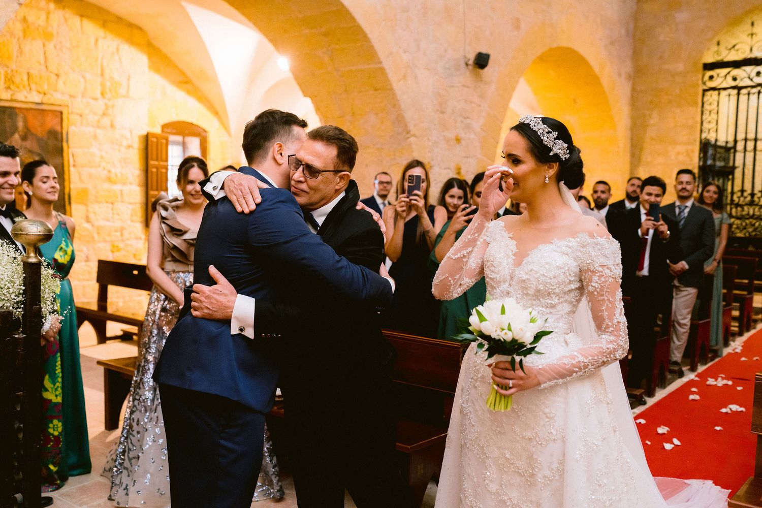 An emotional moment during a wedding ceremony in a stone-walled venue with arched doorways and a red carpet aisle.